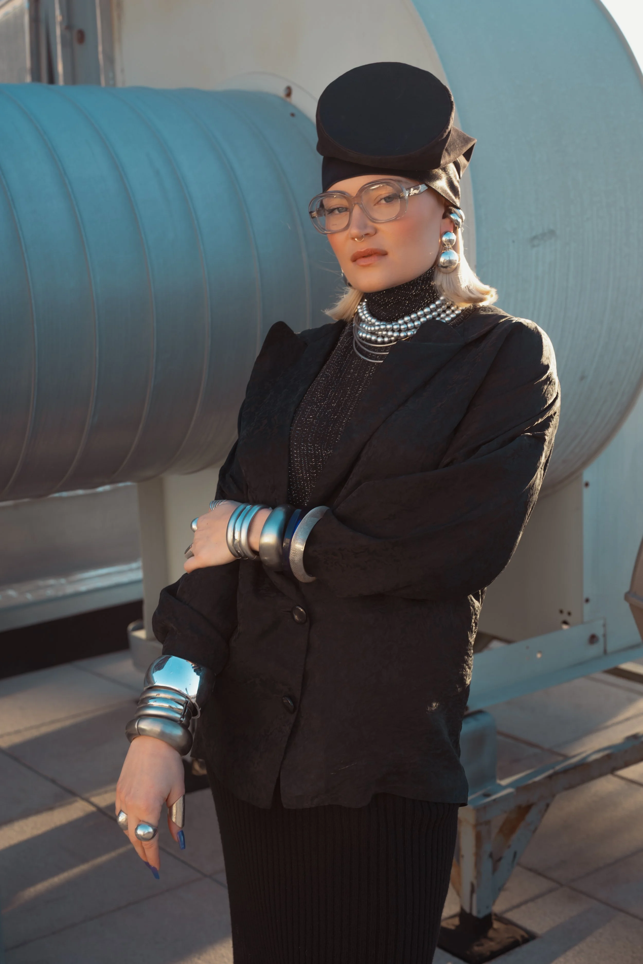 A woman with blonde hair and glasses standing outdoors, wearing a black hat, black jacket, and layered jewelry with large pearl necklaces and bracelets. She is posing with her arms crossed in front of a large metallic industrial structure, possibly o