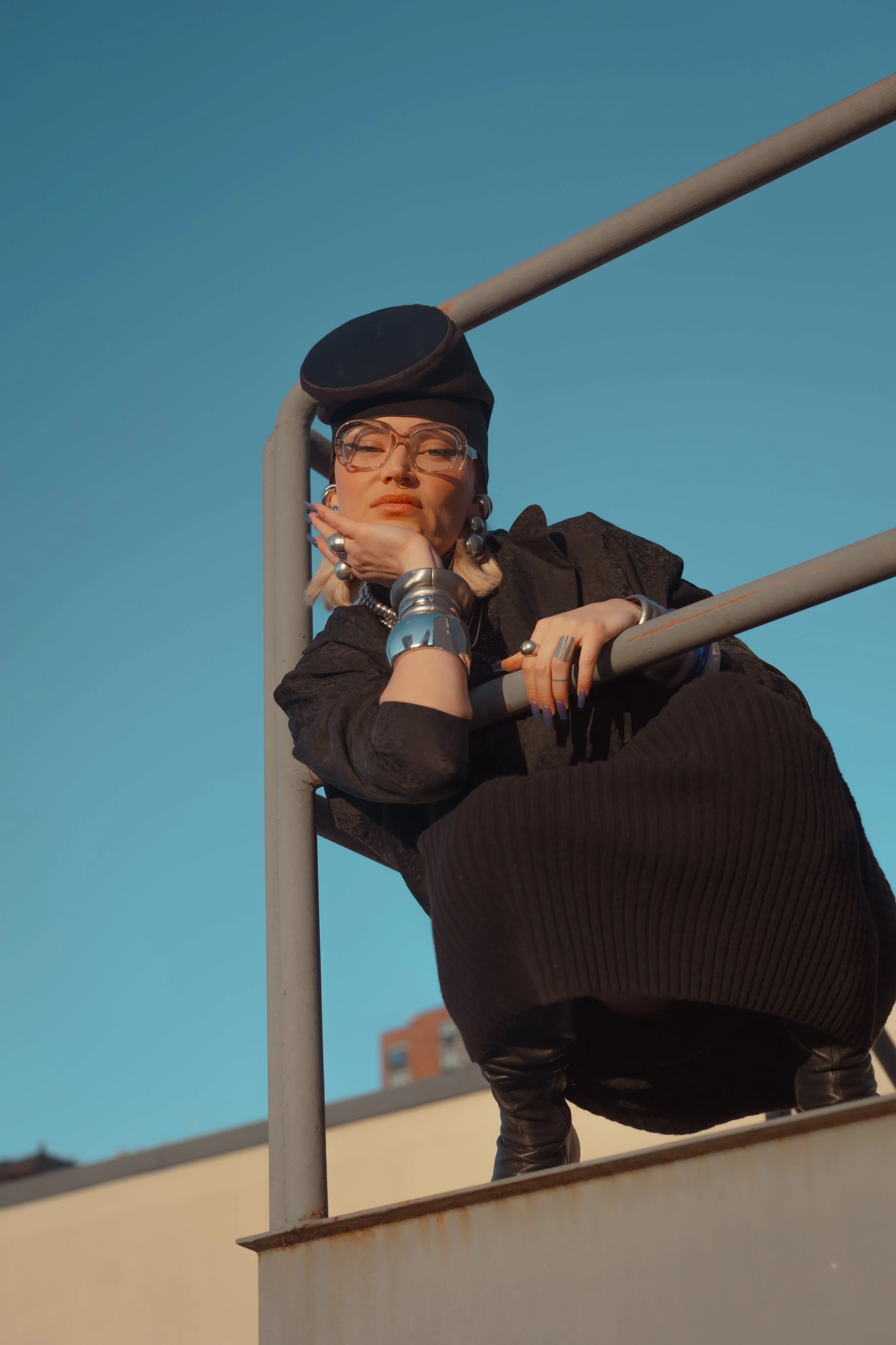 A woman with short blonde hair, glasses, and bold jewelry is leaning on a metal railing, looking at the camera against a blue sky backdrop.