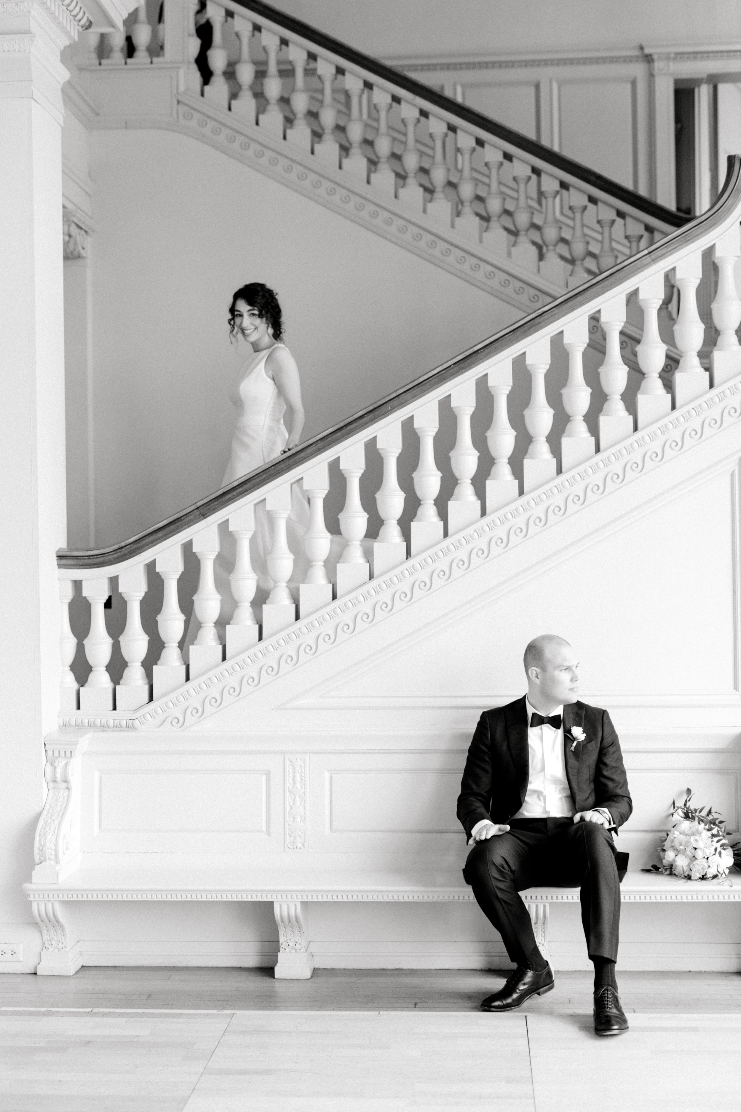 Our bride walks down the grand staircase as her groom waits to see her for the first time for their first look at their romantic and classic wedding day for their french inspired green and white Cairnwood Estate wedding