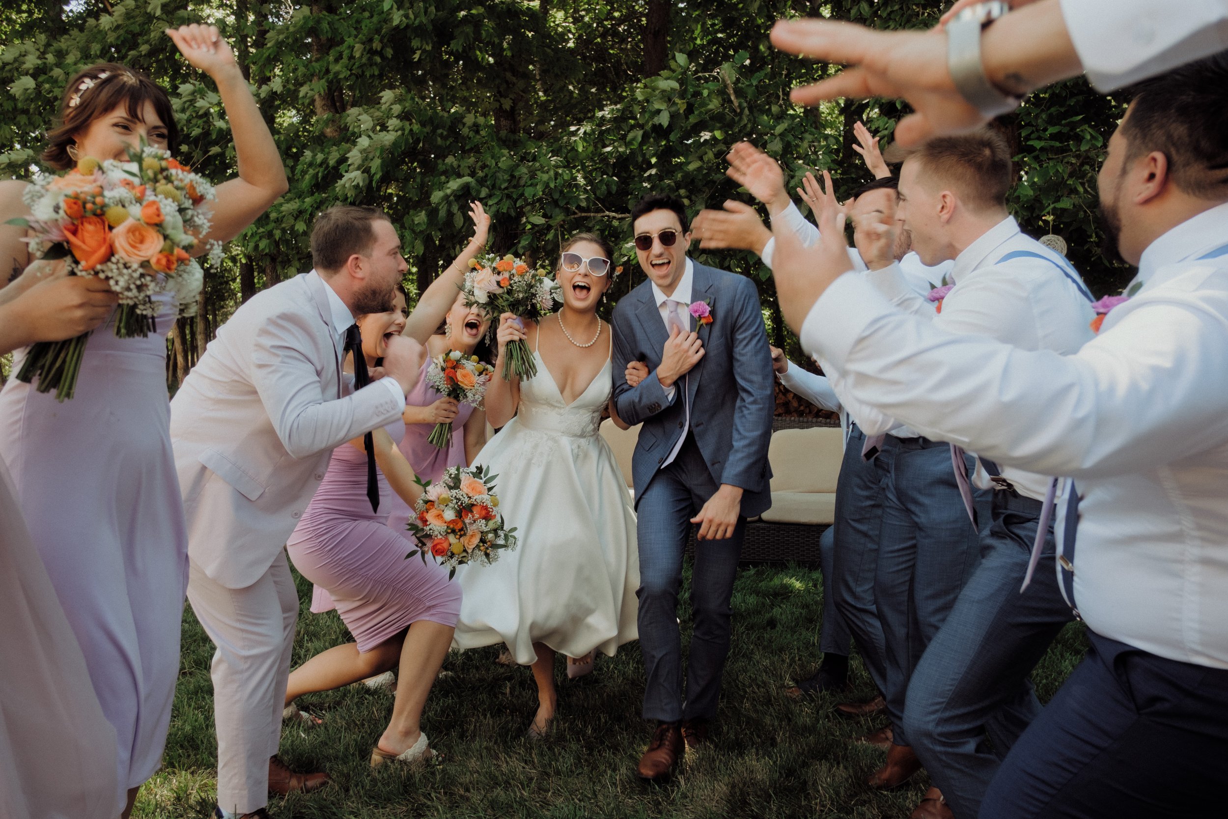 Joyful wedding couple surrounded by cheering bridal party during outdoor garden wedding, candid celebration moment with bouquets and summer greenery.