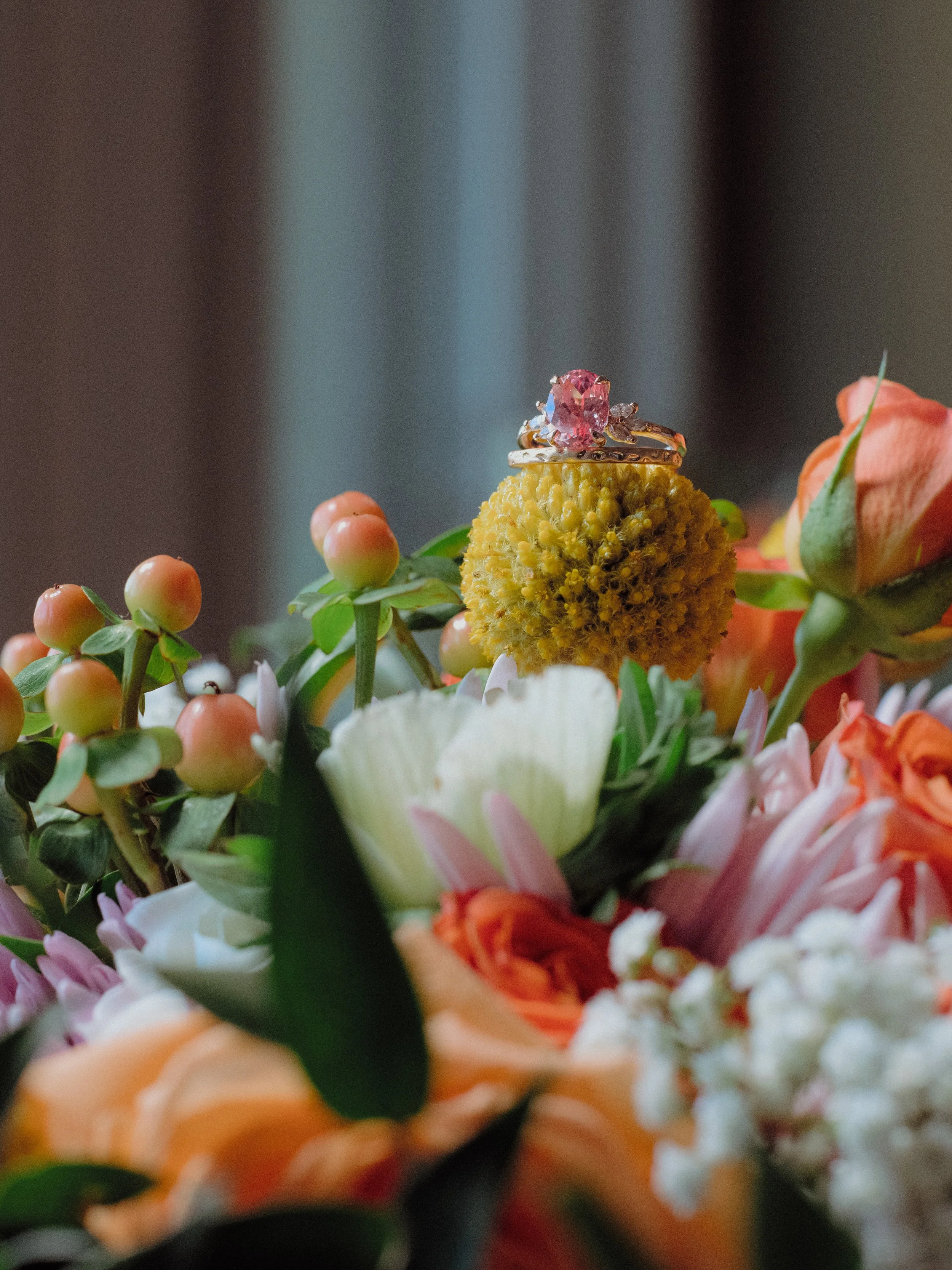 Pink gemstone engagement ring displayed on a yellow billy ball flower surrounded by soft peach and blush wedding florals, romantic close-up detail shot.