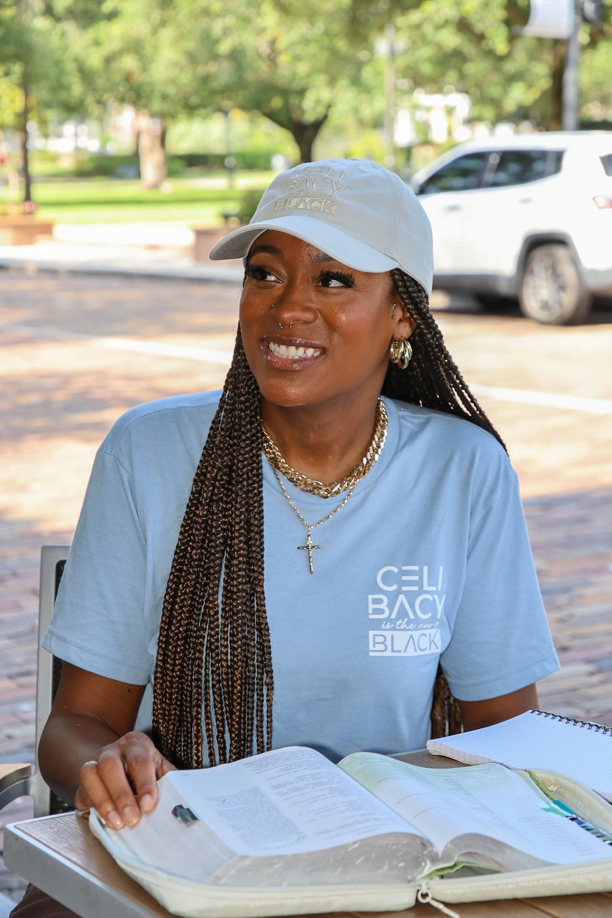 A woman with braided hair wearing a white cap and a light blue T-shirt sitting outdoors at a table with open books, with a background of trees and a white vehicle.