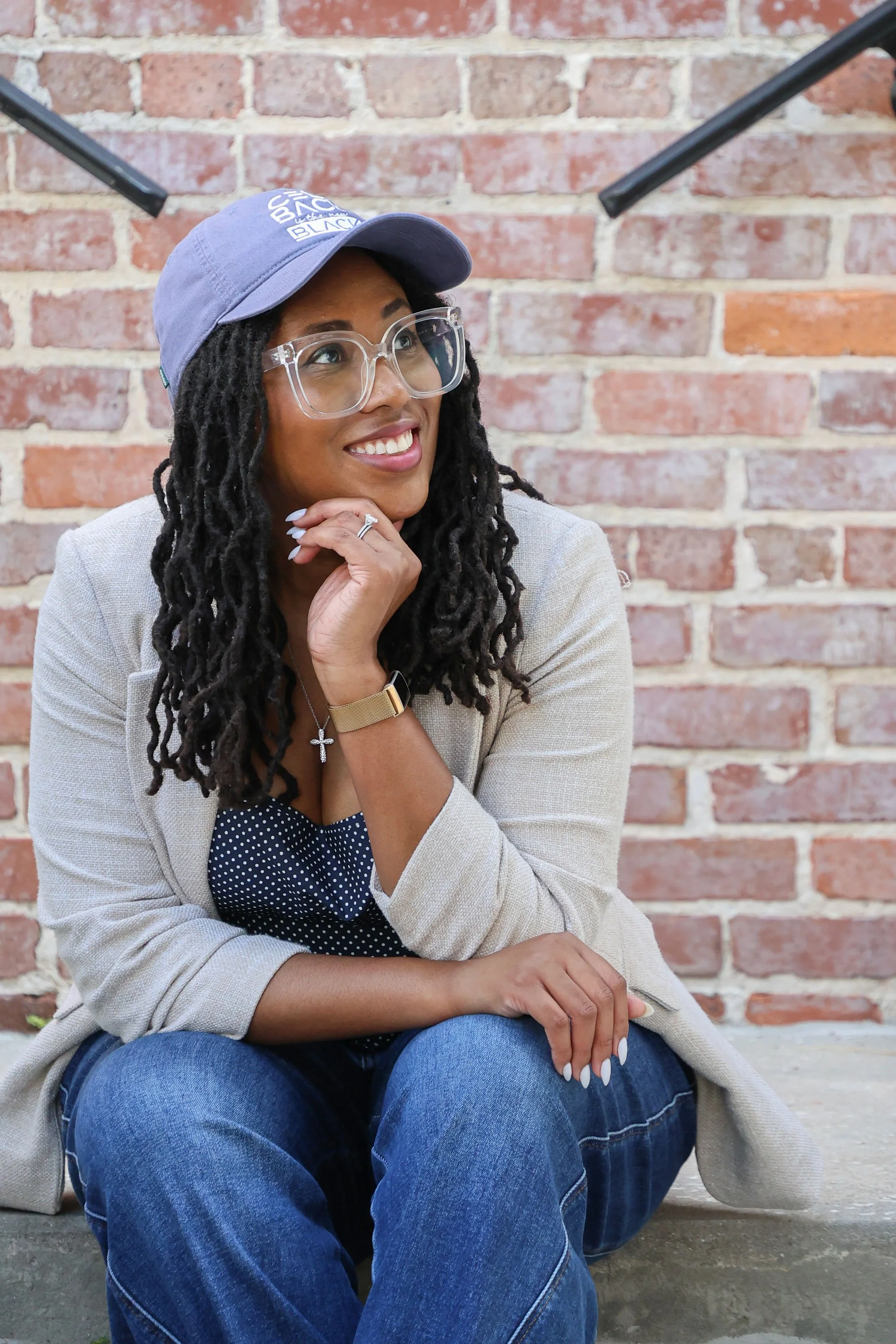 A woman with glasses and dreadlocks, wearing a blue baseball cap, a beige blazer, and jeans, sitting on stairs in front of a red brick wall.