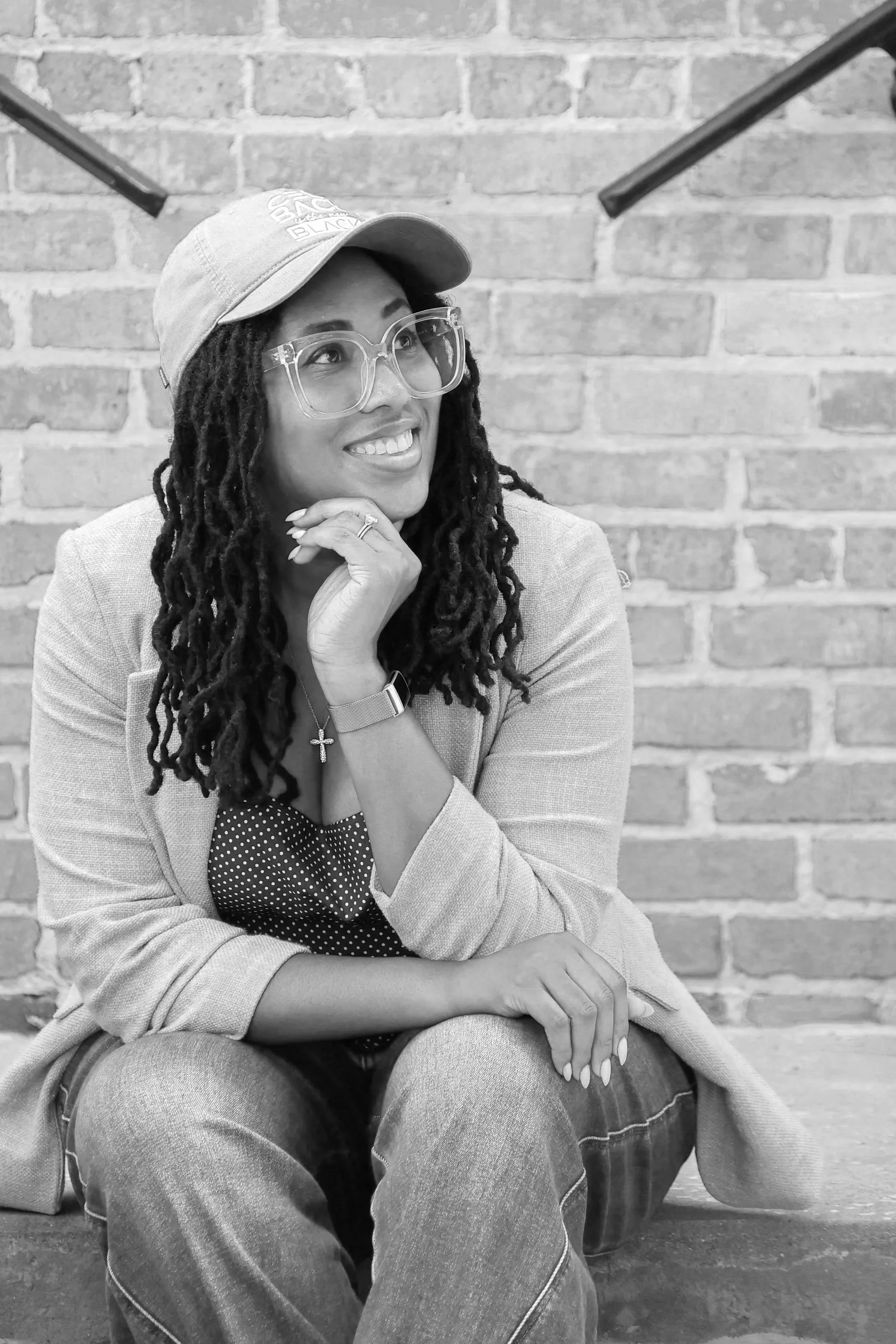 A woman with dreadlocks wearing glasses and a cap, sitting against a brick wall, smiling and resting her chin on her hand.