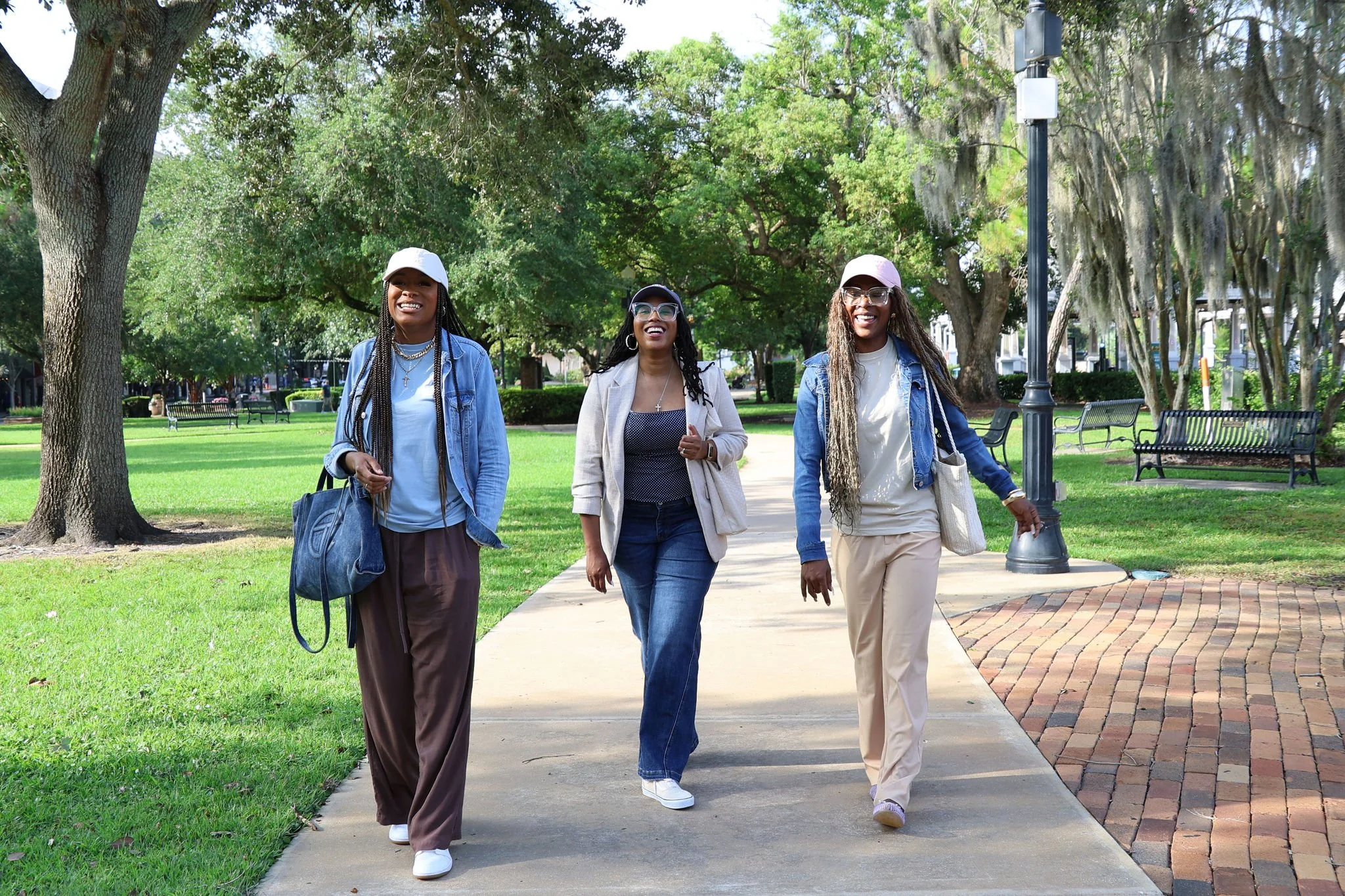 Three women walking and smiling in a park with green trees, benches, and a lamppost.