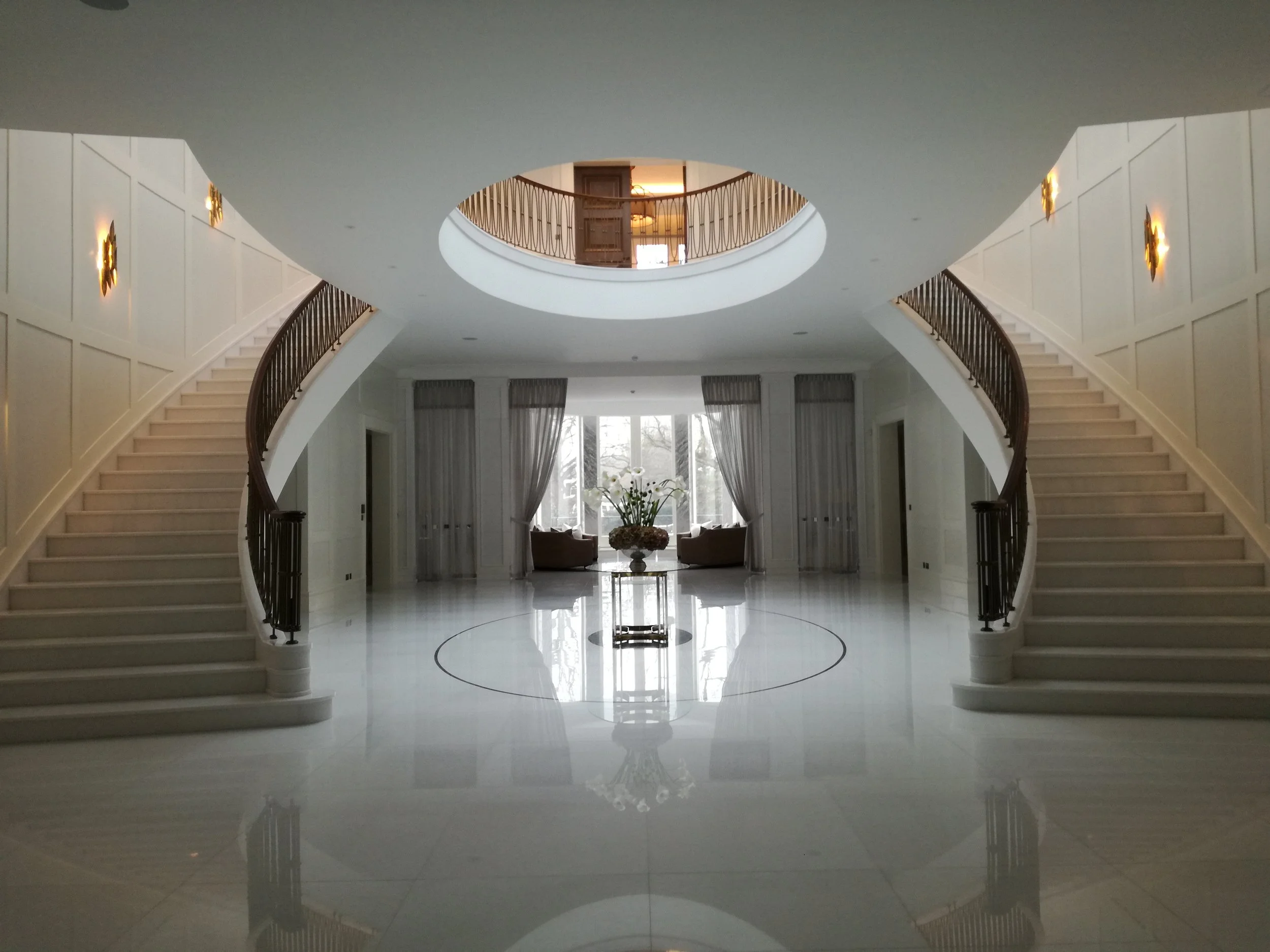 Elegant hotel lobby with two symmetrical curved staircases, large windows with curtains, and a central floral arrangement. The floor is polished and reflective.