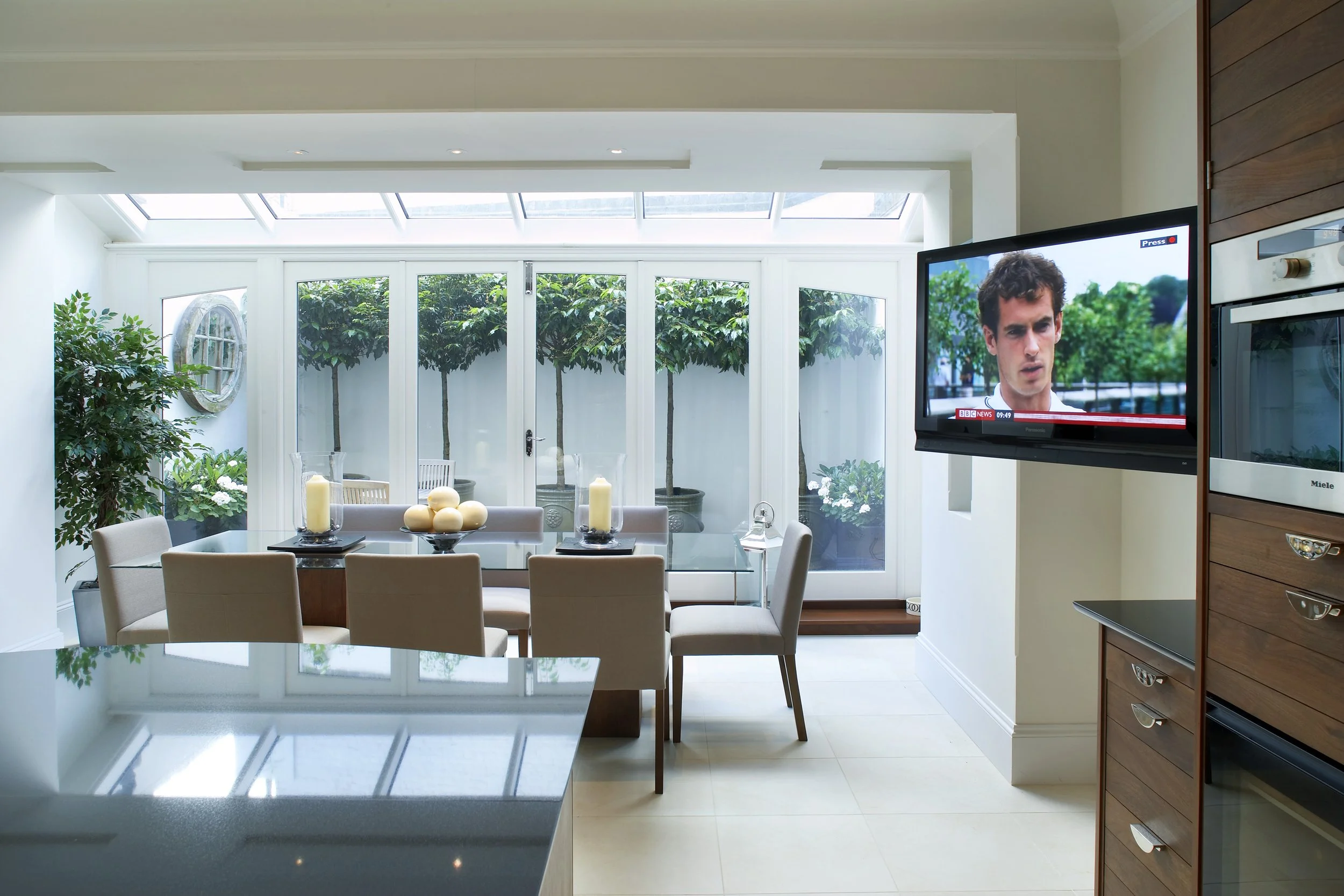 Bright kitchen and dining area with a glass table, seven beige chairs, candles in glass holders, a kitchen wardrobe, and a mounted TV showing a news program, with large windows and potted trees outside.