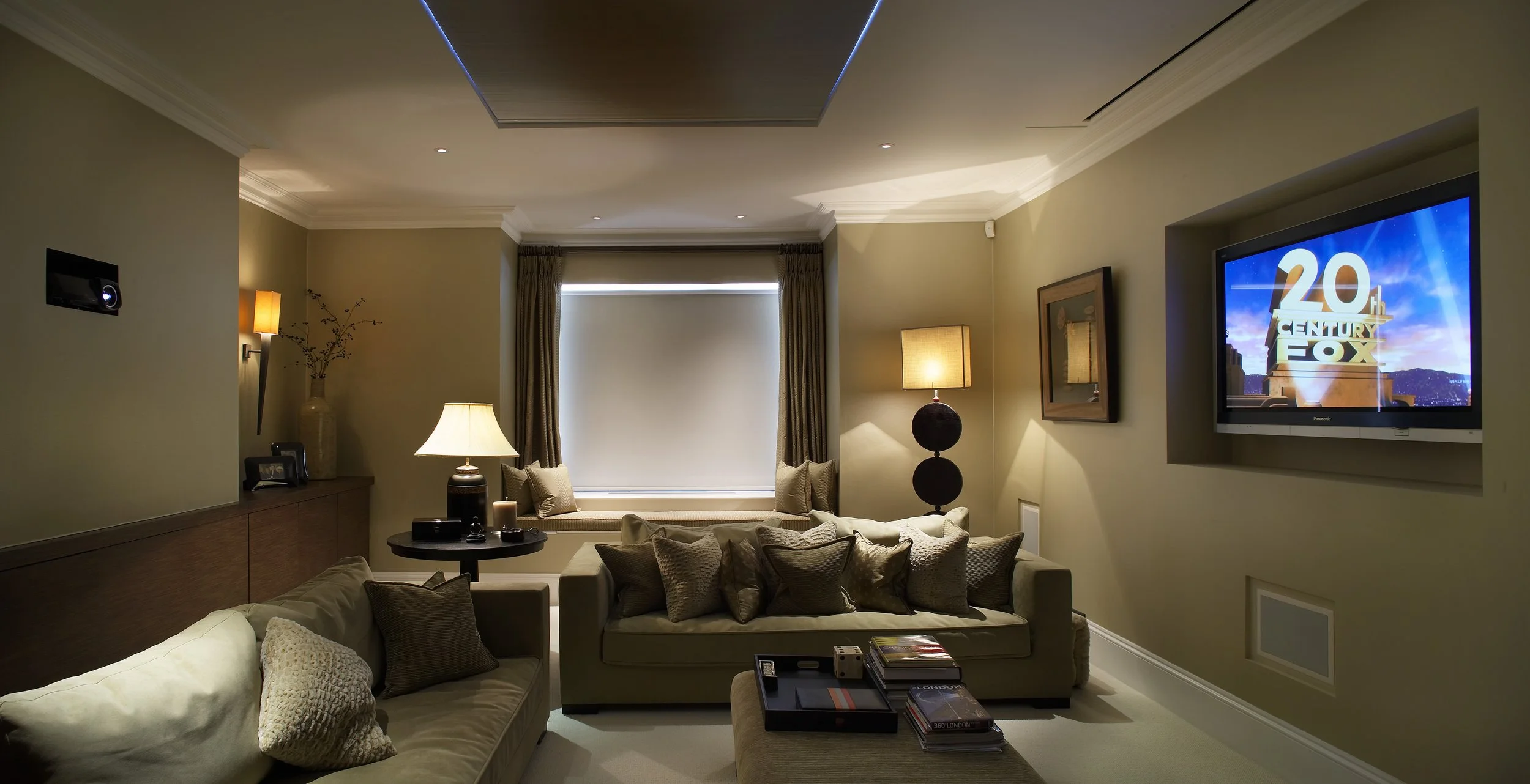 A living room with beige walls, a flat-screen TV displaying the 20th Century Fox logo, beige sofas with numerous pillows, a large window with curtains, a small round table with lamps, and a wall-mounted speaker.