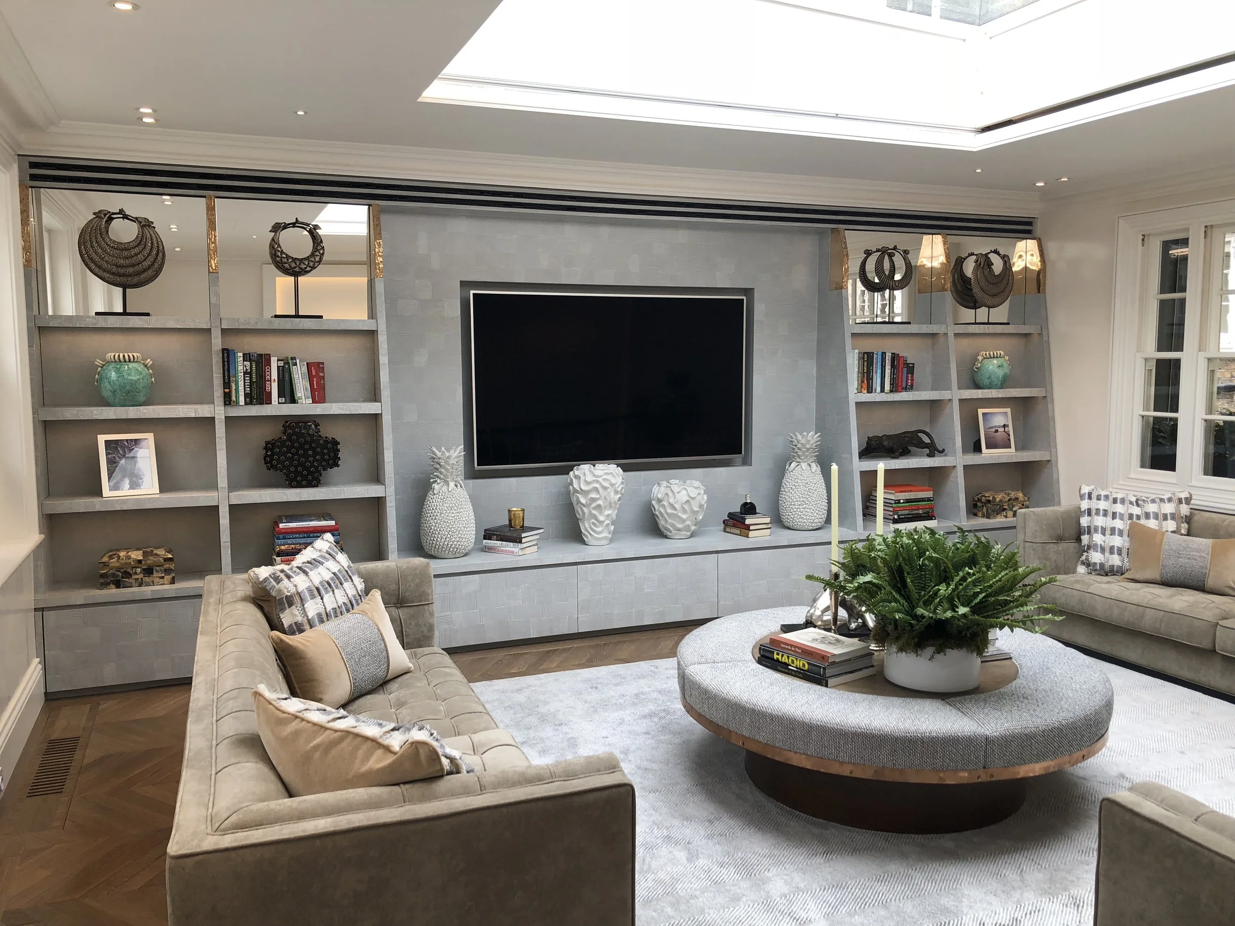 Living room with built-in shelves, a wall-mounted TV, decorative vases, books, a large round coffee table with plants and candlesticks, beige sofas with pillows, and a window on the right side.