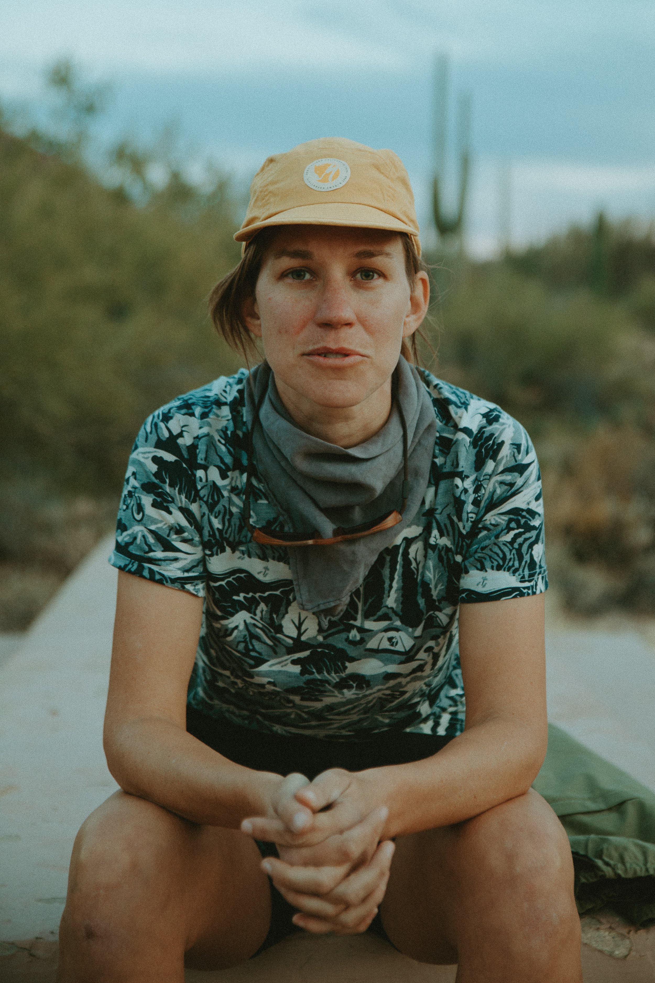 A woman sitting outdoors on a sandy surface, wearing a beige cap, a patterned shirt, and a neck gaiter, with desert vegetation and cacti in the background.