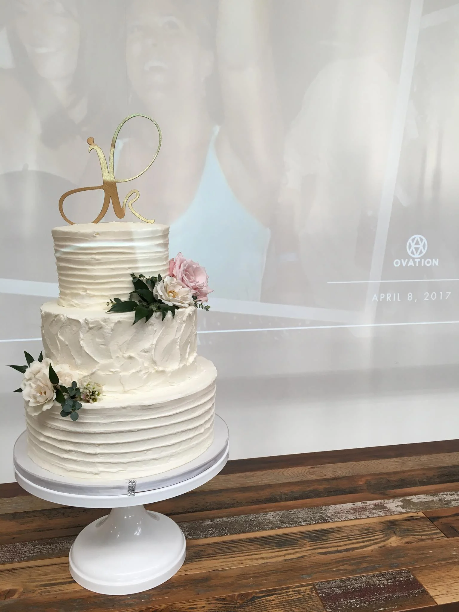 Three-tier wedding cake with white frosting, decorated with pink and white flowers, and topped with a gold cake topper shaped as initials.