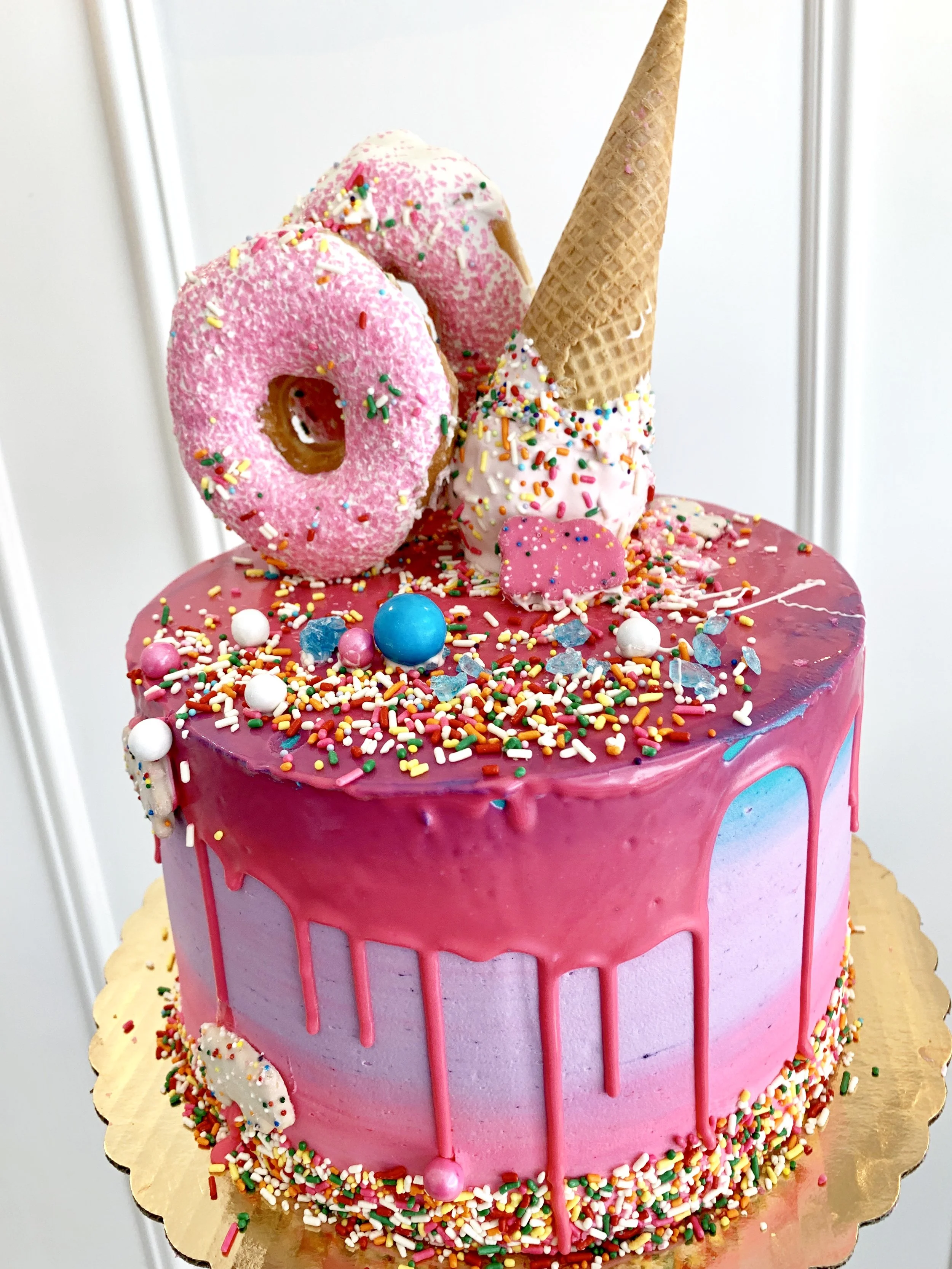 A colorful birthday cake with pink glaze, rainbow sprinkles, cookies, ice cream cones, and edible decorations.
