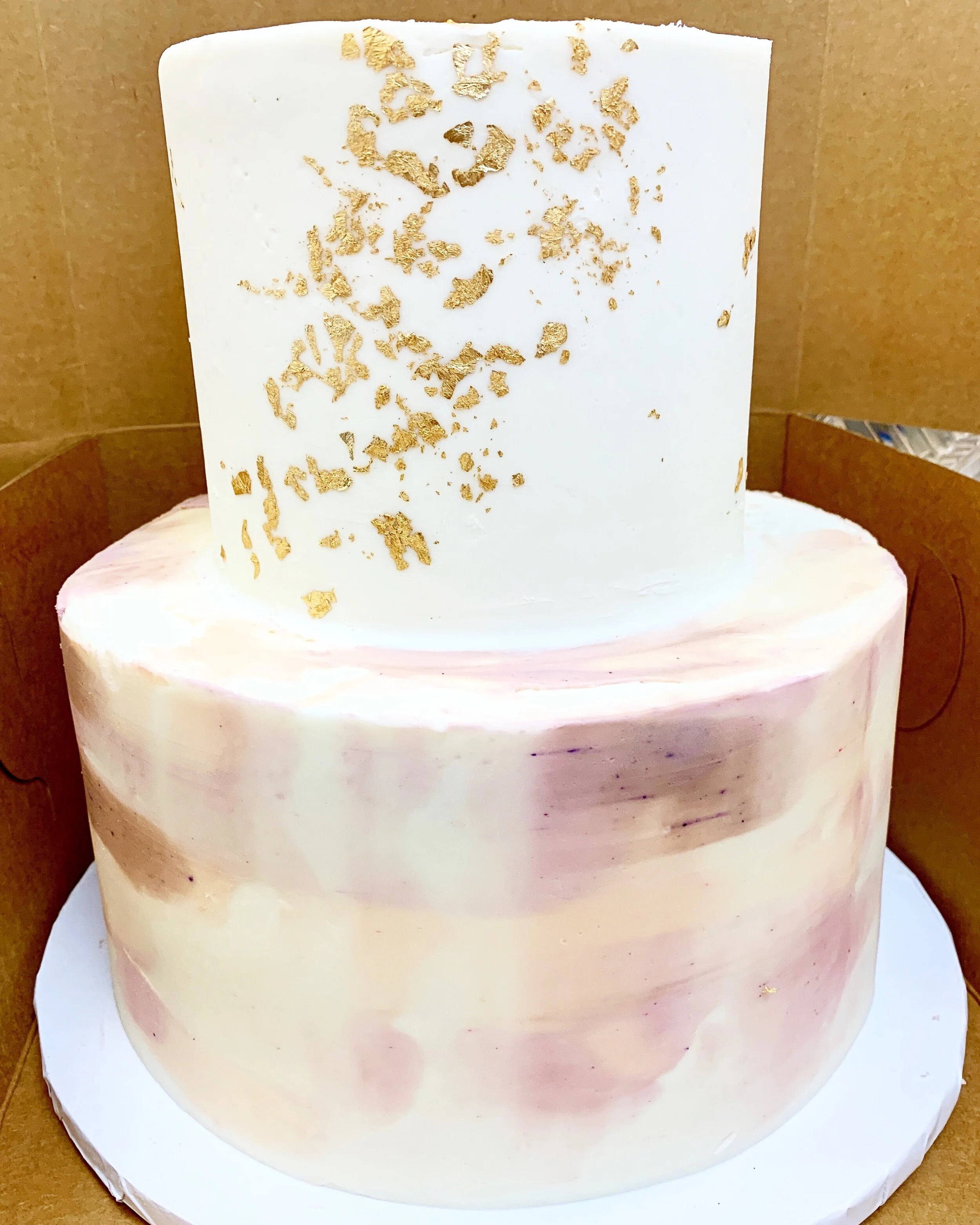 A two-tiered cake with white and pink marbled buttercream, adorned with gold leaf on the top tier.