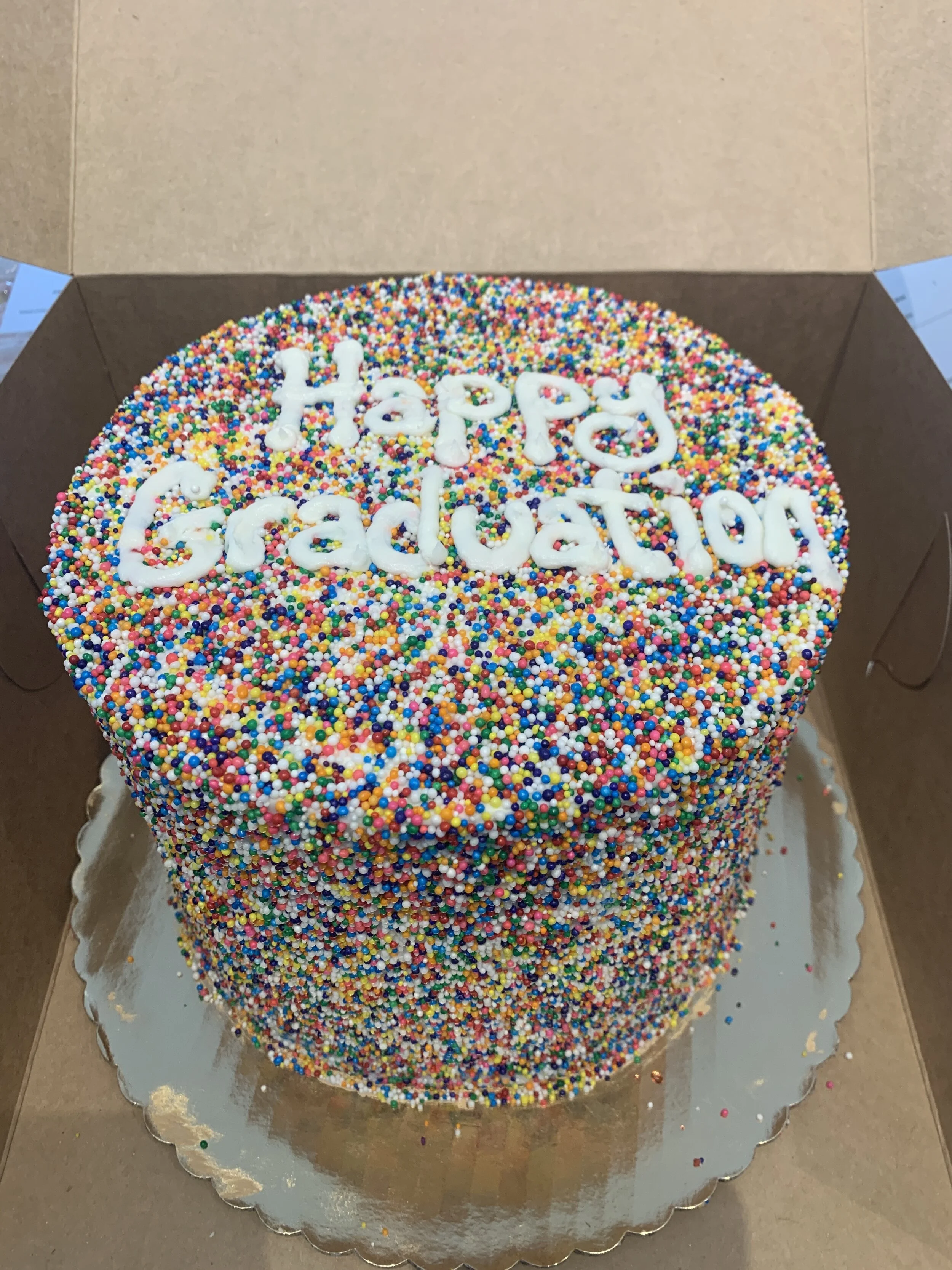 A colorful round cake decorated with multicolored sprinkles and white icing that says 'Happy Graduation' on top.