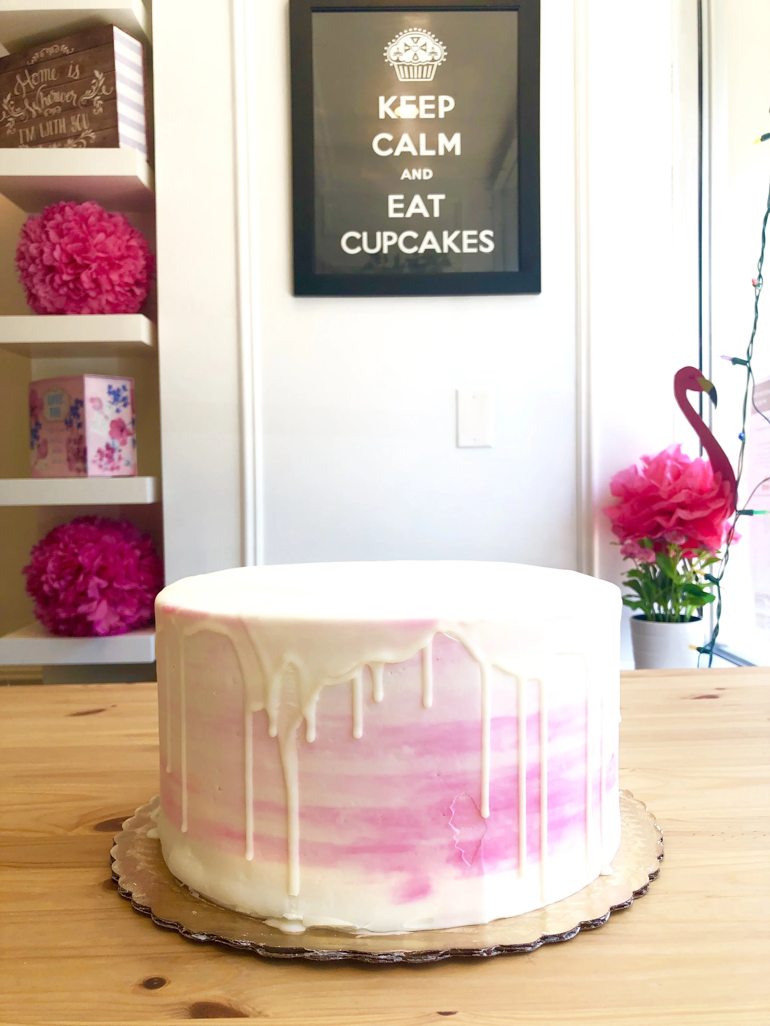 A round cake with pink and white marbled frosting and white drip decoration on a wooden table. The background has a black sign with white text that says "Keep calm and eat cupcakes", pink pom-poms, a potted pink flower, and a flamingo decoration.