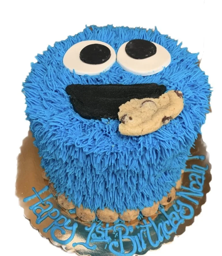 A birthday cake designed to look like Cookie Monster from Sesame Street, covered in blue frosting with piped fur texture, large googly eyes, a black mouth, and a cookie on his tongue, on a gold board with 'Happy 1st Birthday' written in blue icing.