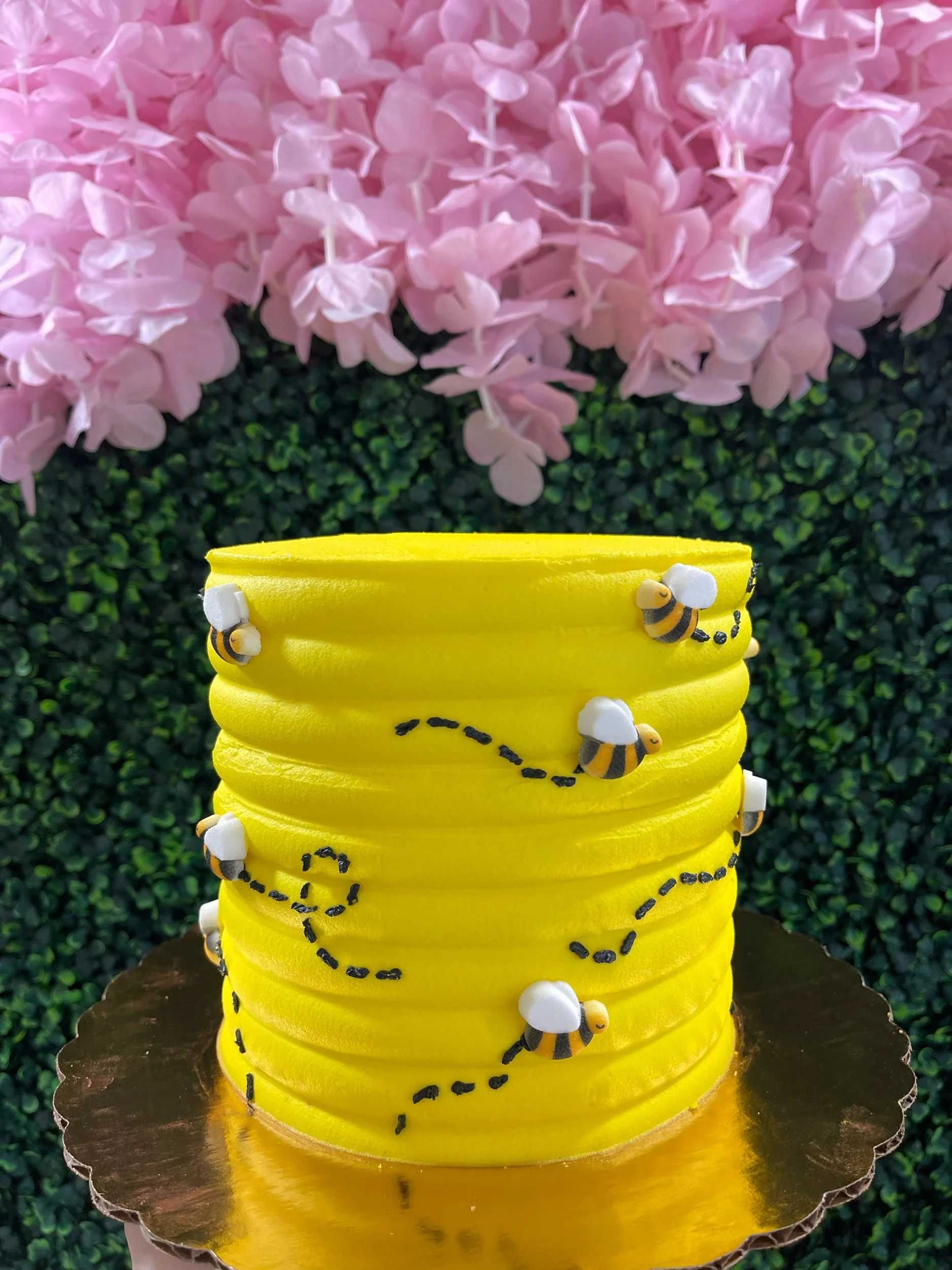 Yellow cake decorated with bee figurines and black dotted lines resembling bee flight paths, set on a golden cake board, with pink flowers and green foliage in the background.