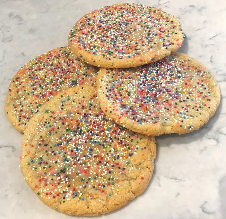 Four sugar cookies decorated with colorful rainbow sprinkles on a marble surface.