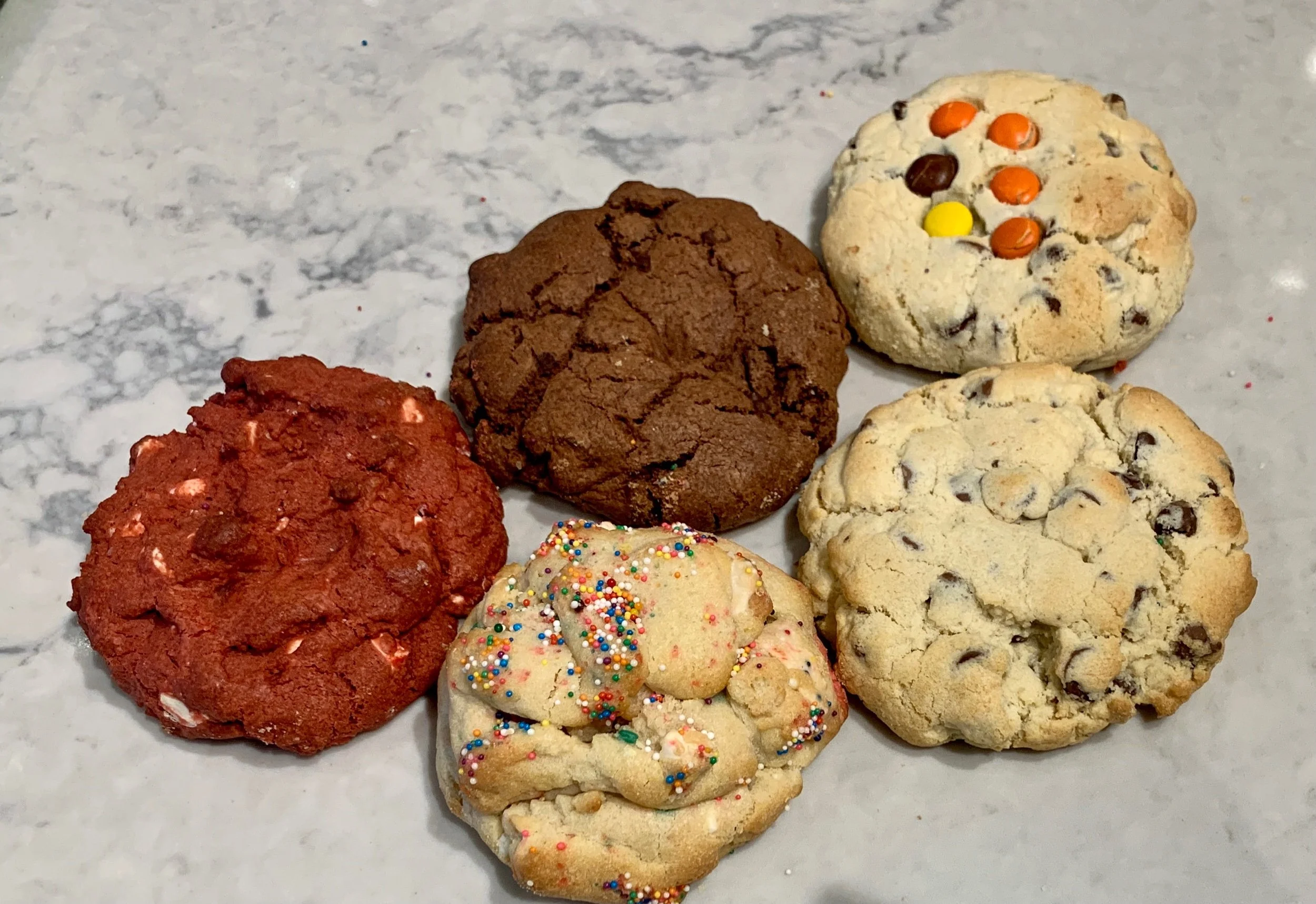 Six assorted cookies on a marble surface, including a red velvet cookie with white chocolate chips, a double chocolate cookie, a sugar cookie with rainbow sprinkles, and three chocolate chip cookies.