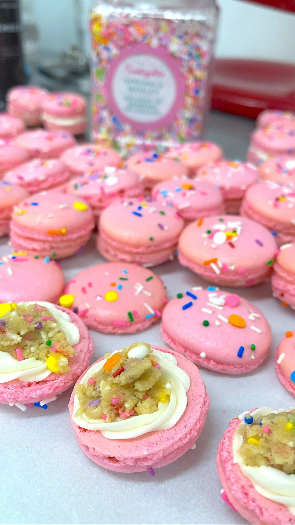 Pink macarons with colorful sprinkles, some with cream and sprinkles as filling, on a white surface with a box of sprinkles in the background.