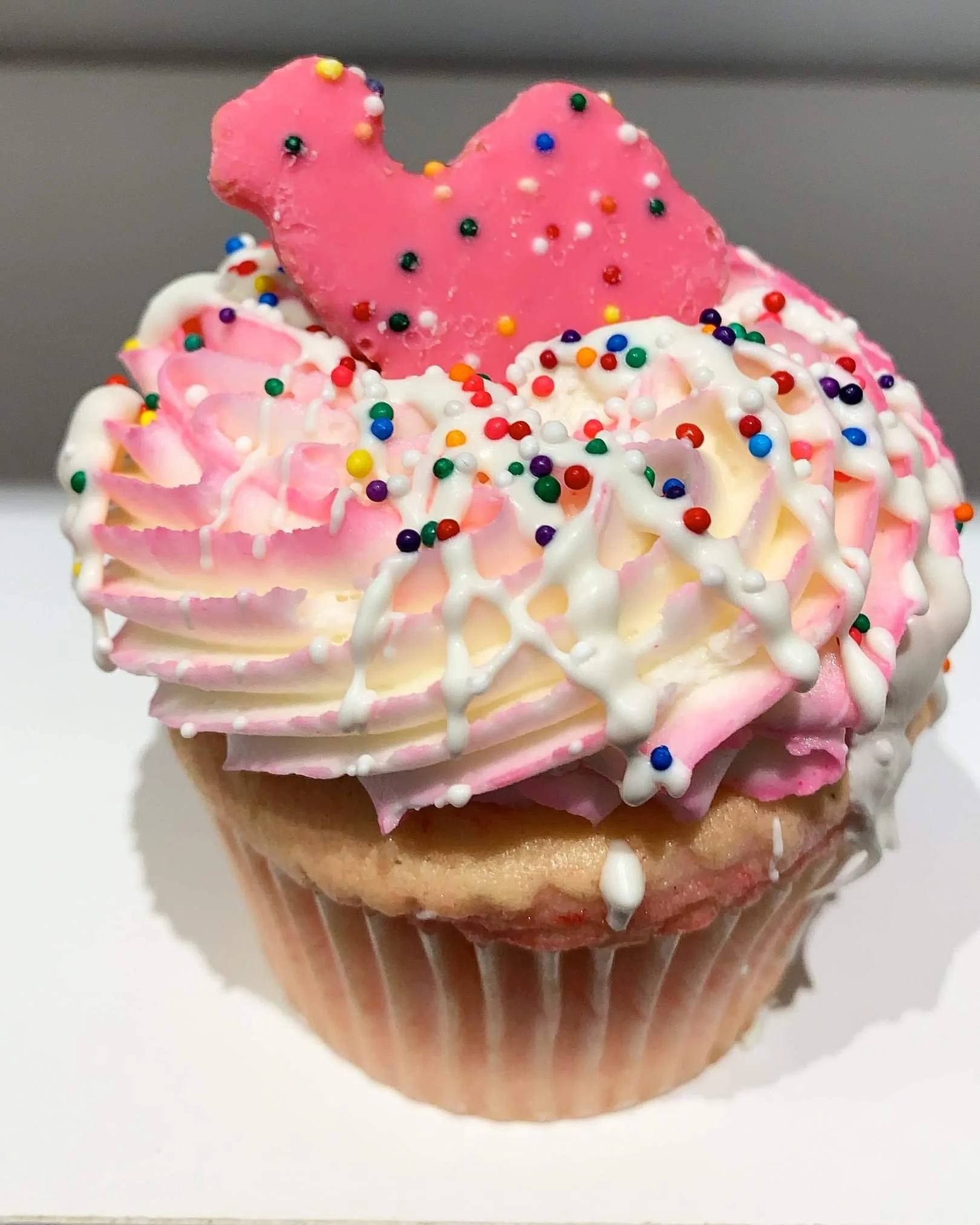 Cupcake with pink frosting, colorful sprinkles, white drizzle, and a pink heart-shaped cookie on top.