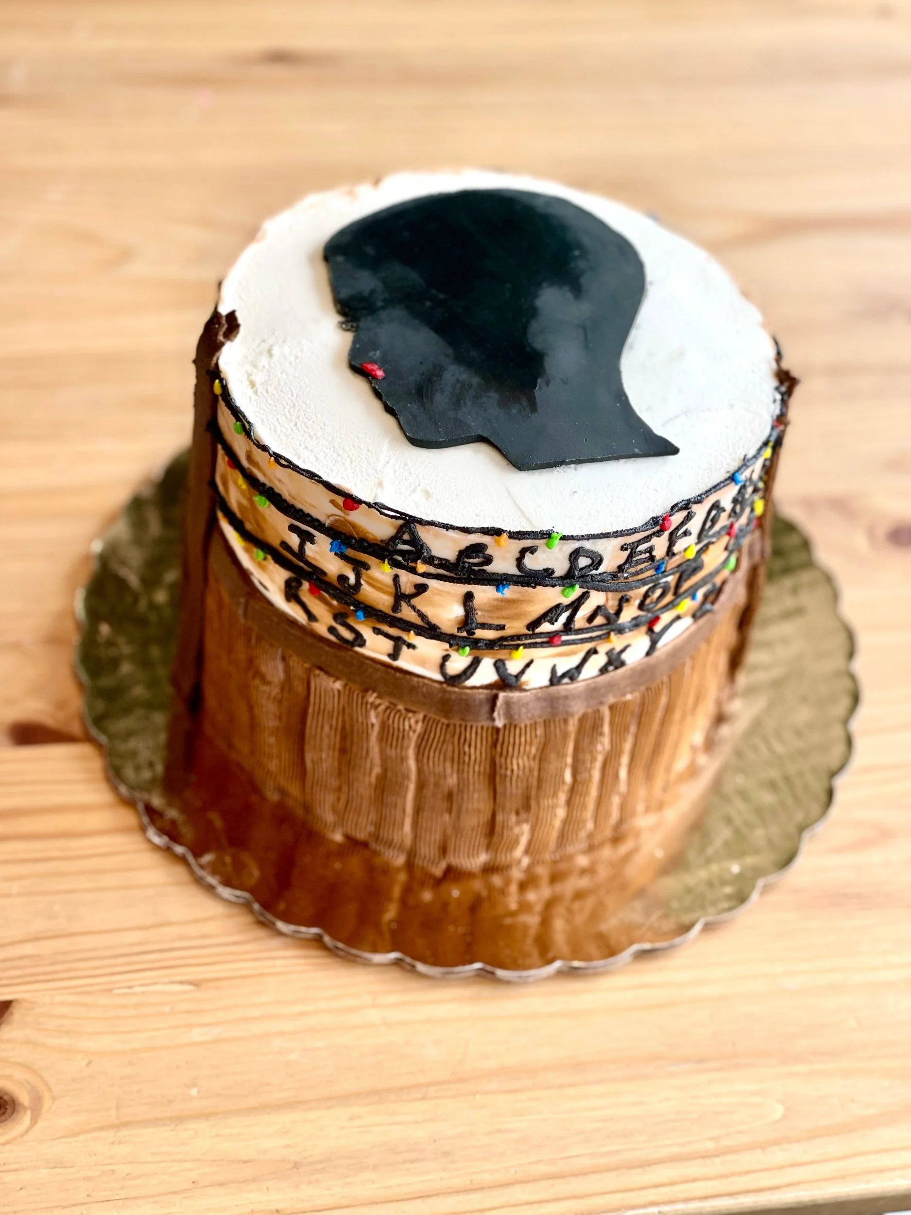 A birthday cake designed to look like a tree trunk with a chocolate top featuring a black silhouette of a person's profile and alphabet letters around the side, decorated with multicolored sprinkles.
