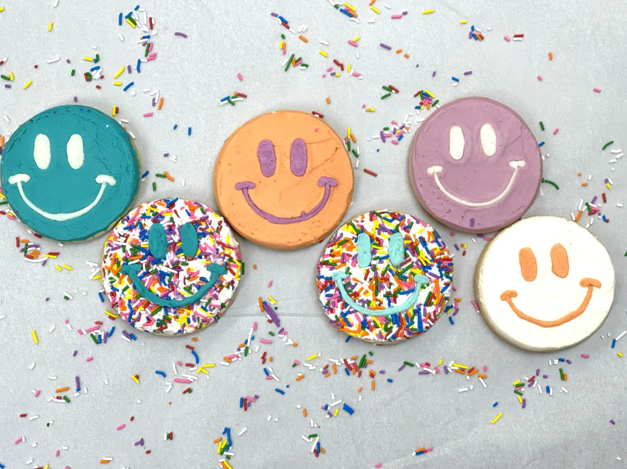 Colorful decorated cookies with smiley faces, surrounded by rainbow sprinkles on a white surface.
