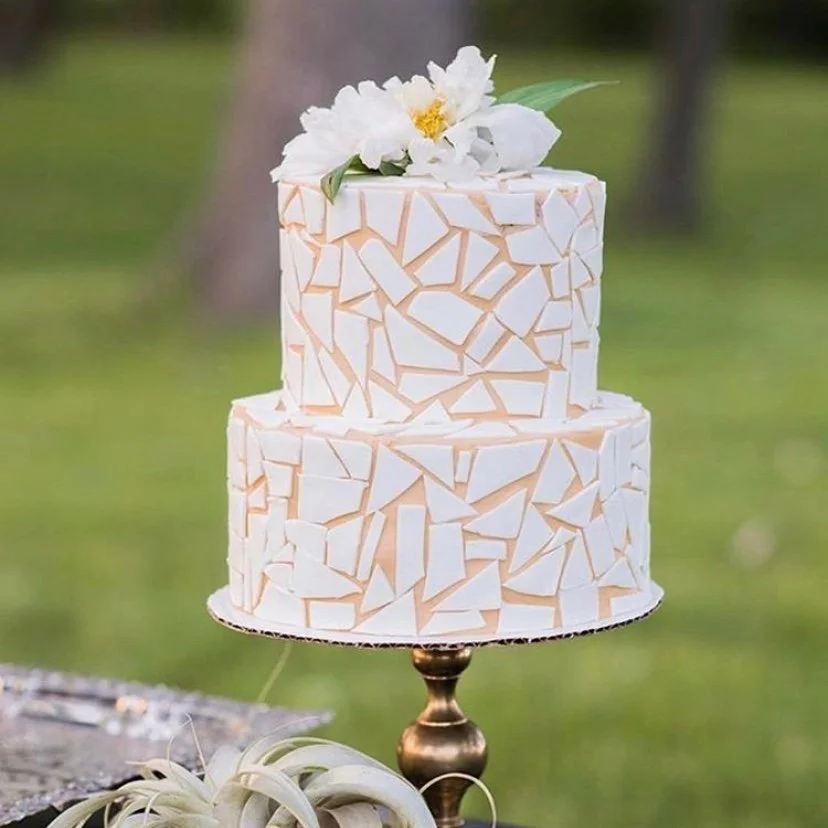 A two-tier wedding cake decorated with irregular white and beige mosaic patterns, topped with white flowers and green leaves, placed on a gold cake stand outdoors.