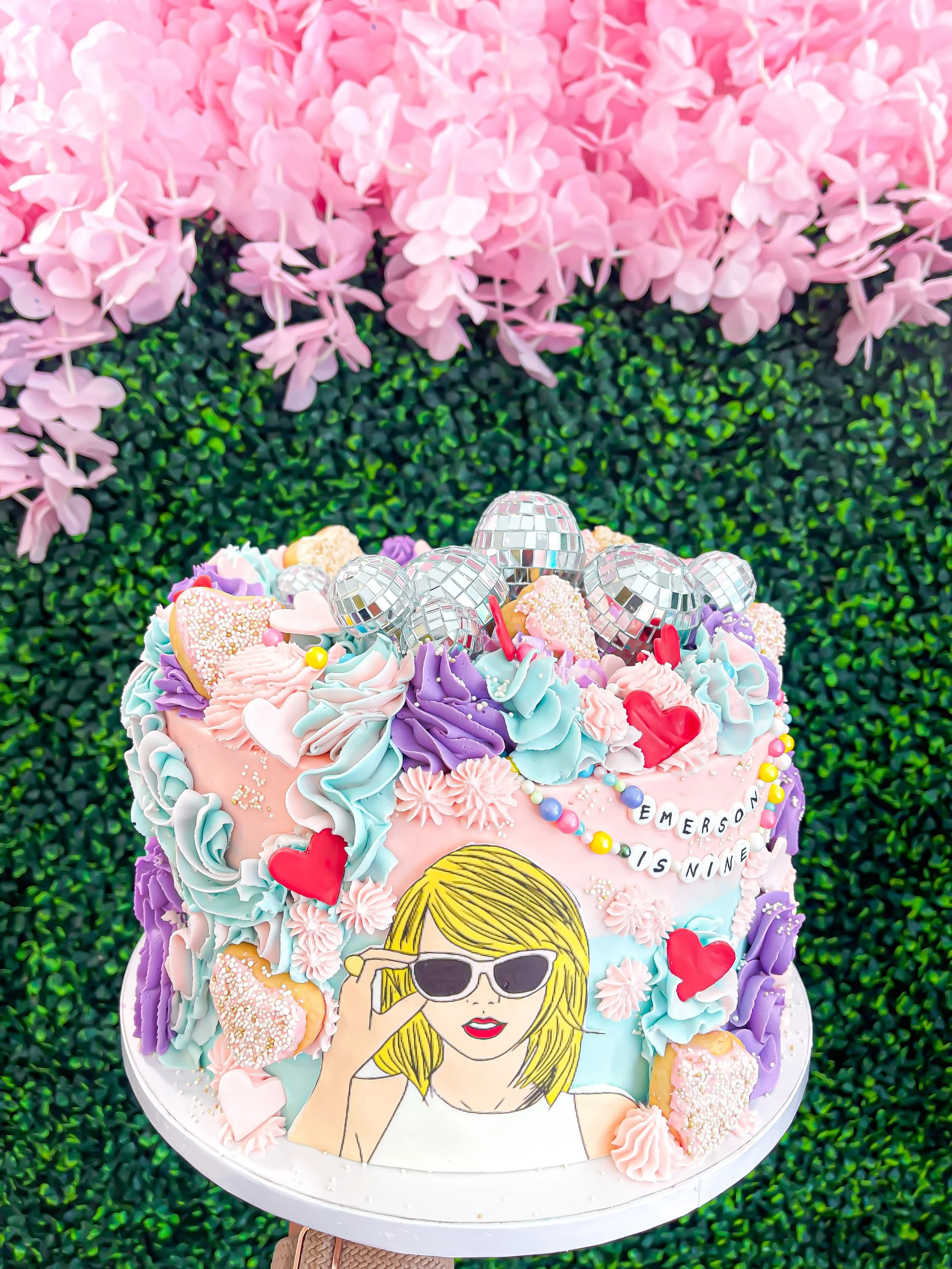 Colorful birthday cake decorated with pastel-colored frosting, heart-shaped cookies, disco balls, and a cartoon image of a girl with blonde hair and sunglasses, placed on a white cake stand with pink flowers in the background.
