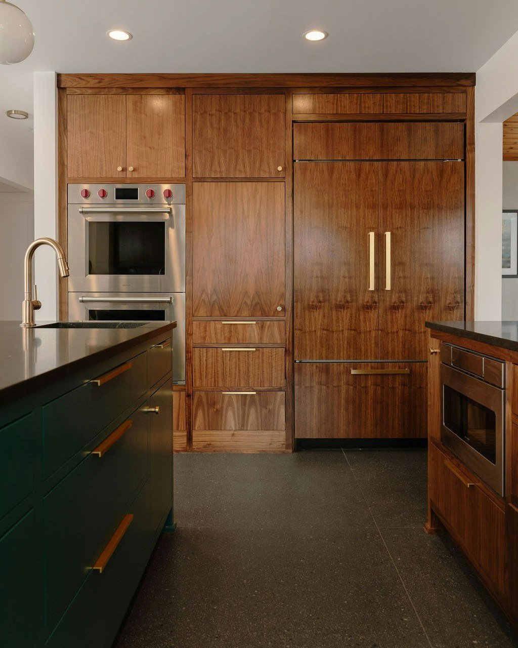 A floor to ceiling interior wall offers optimal space for positioning the trio of tall kitchen essentials: double ovens, pantry, refrigerator. Cladding the refrigerator with coordinating walnut panels seamlessly integrates functionality with a striki