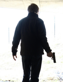 Person walking outdoors holding a handgun in their right hand, viewed from behind.
