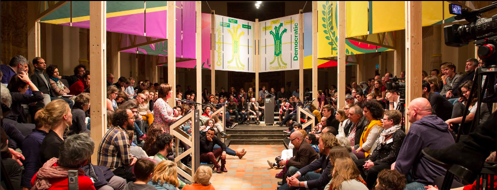 "Ik wil geen kunst maken in een zogeheten democratie. Ik wil de democratie mede vormgeven. (uit: Kunst ter verdediging van de democratie, Jonas Staal, 2012) Jonas Staal is de oprichter van New World Summit een artistieke en politieke organisatie die zich bezig houdt met het organiseren van alternatieve parlementen. Alternatief omdat het parlement bestaat uit groeperingen die worden uitgesloten van democratie. Jonas nodigde in het verleden onder andere de Koerdische vrouwenbeweging uit Rojava uit (noorden van Syrië), waar in 2012 een stateloze democratie is uitgeroepen op basis van principes als zelfbestuur en gelijkheid van gender. Naast de politieke inhoud van zijn project, hecht hij ook veel belang aan de vormgeving van deze pop – up parlementen. Welke impact heeft de vorm van het parlement, cirkelvormig of rechthoekig, op de communicatie tussen de diverse vertegenwoordigers? Wat is het verschil tussen een bijeenkomst in open lucht of in een gesloten ruimte, als de deelnemers op stoelen plaatsnemen of op lange banken?