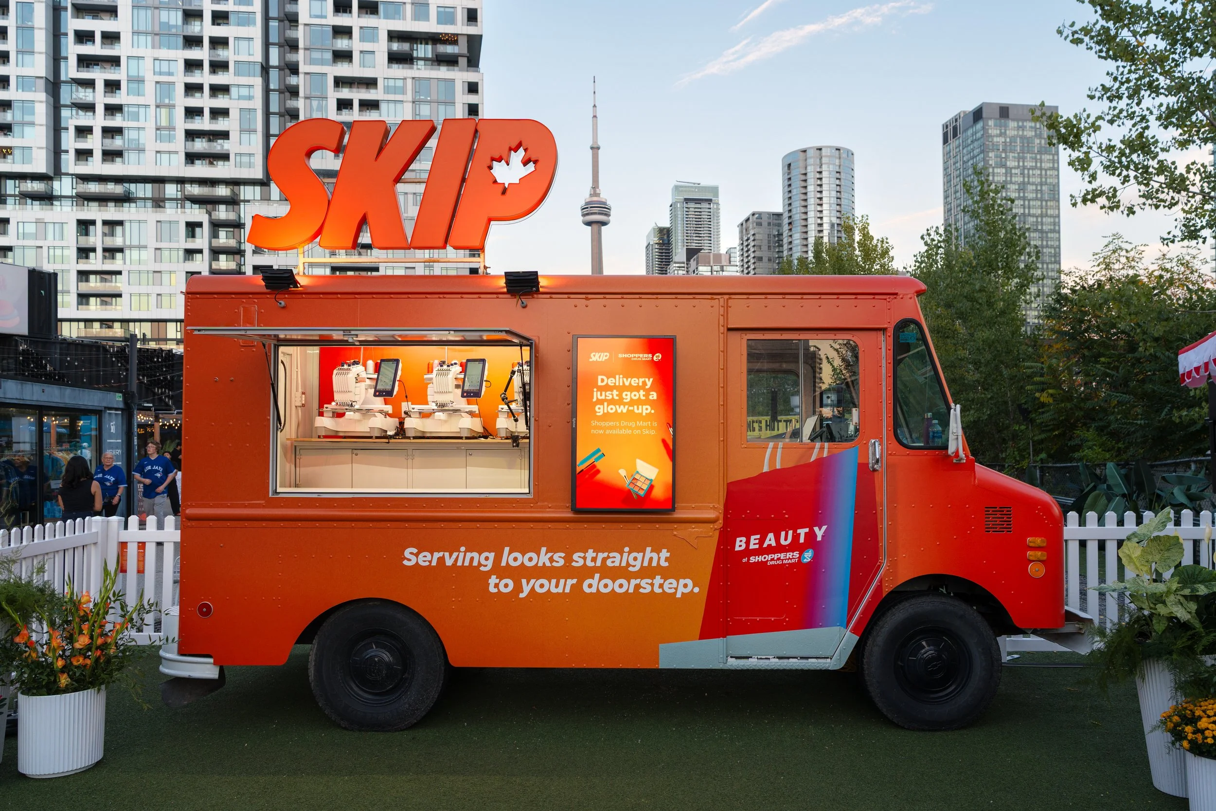 An orange food truck with the words 'Serving looks straight to your doorstep' parked outdoors in front of a city skyline with tall buildings and the CN Tower in the background. The truck has a large 'SKIP' sign with a maple leaf inside the 'I'. There are people standing nearby, and potted plants are around the truck.