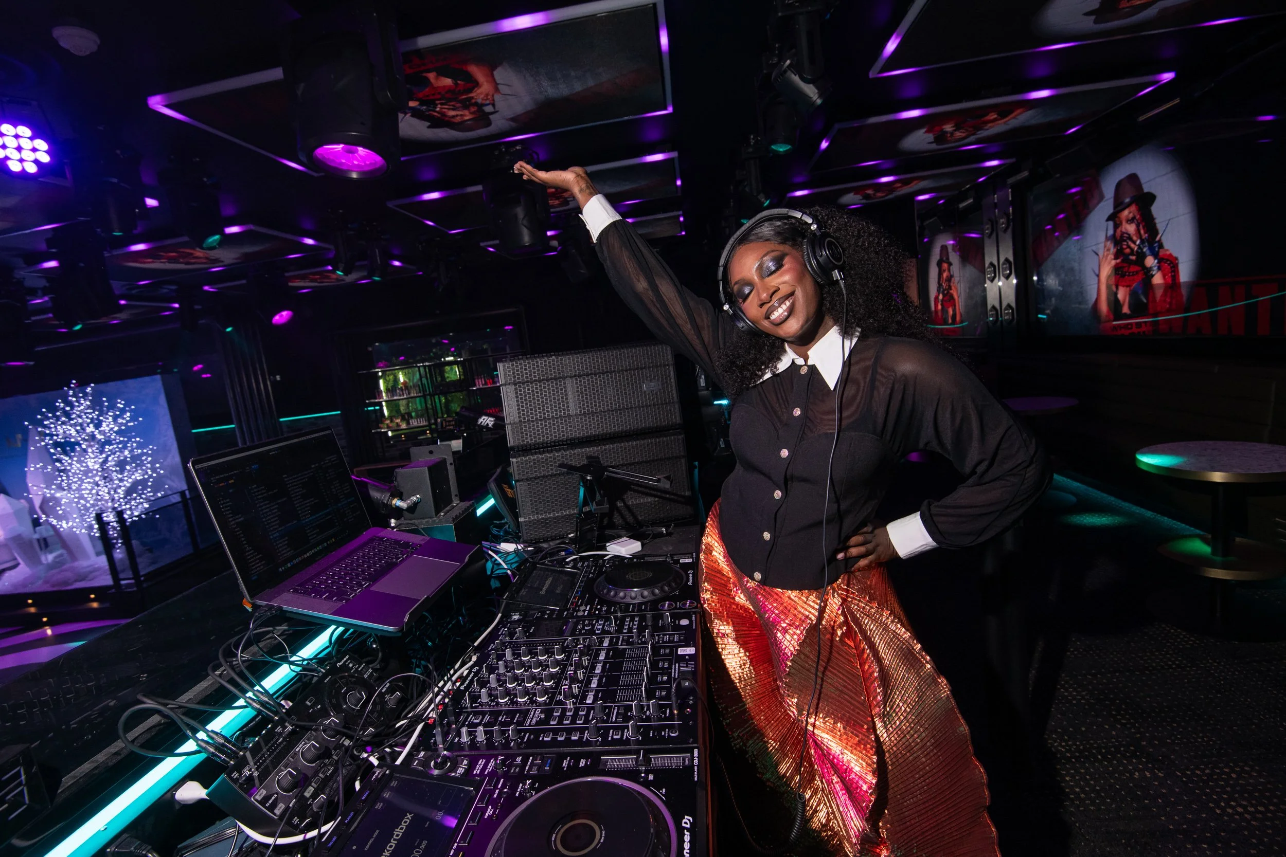 A woman DJ lissa Monet in a nightclub with purple and blue lights, smiling and dancing in front of DJ equipment, wearing headphones, a black shirt with white collar, and shiny orange and pink pants.