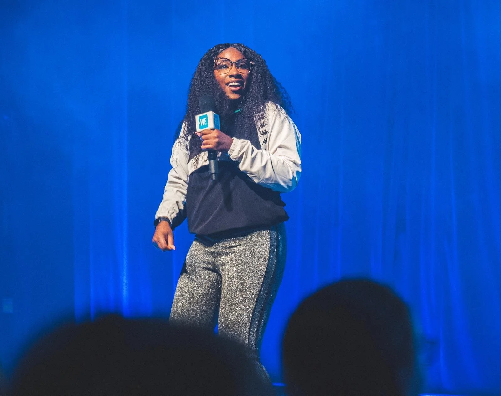 A woman standing on stage holding a microphone with a We logo, wearing glasses, a light jacket, and glittery leggings, smiling with a blue curtain background.