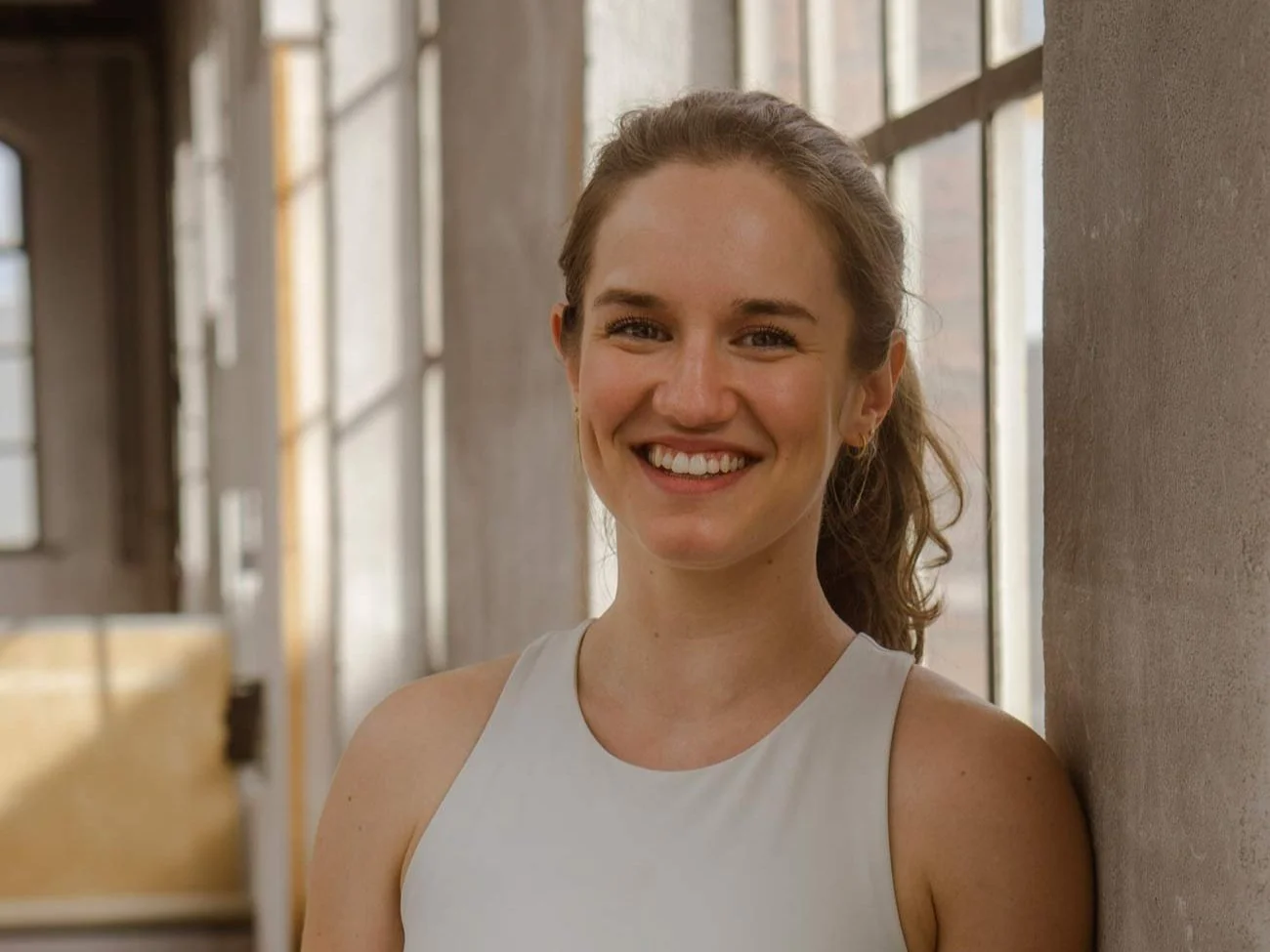 Frau steht lächelnd in einem sonnigen Raum mit großen Fenstern.