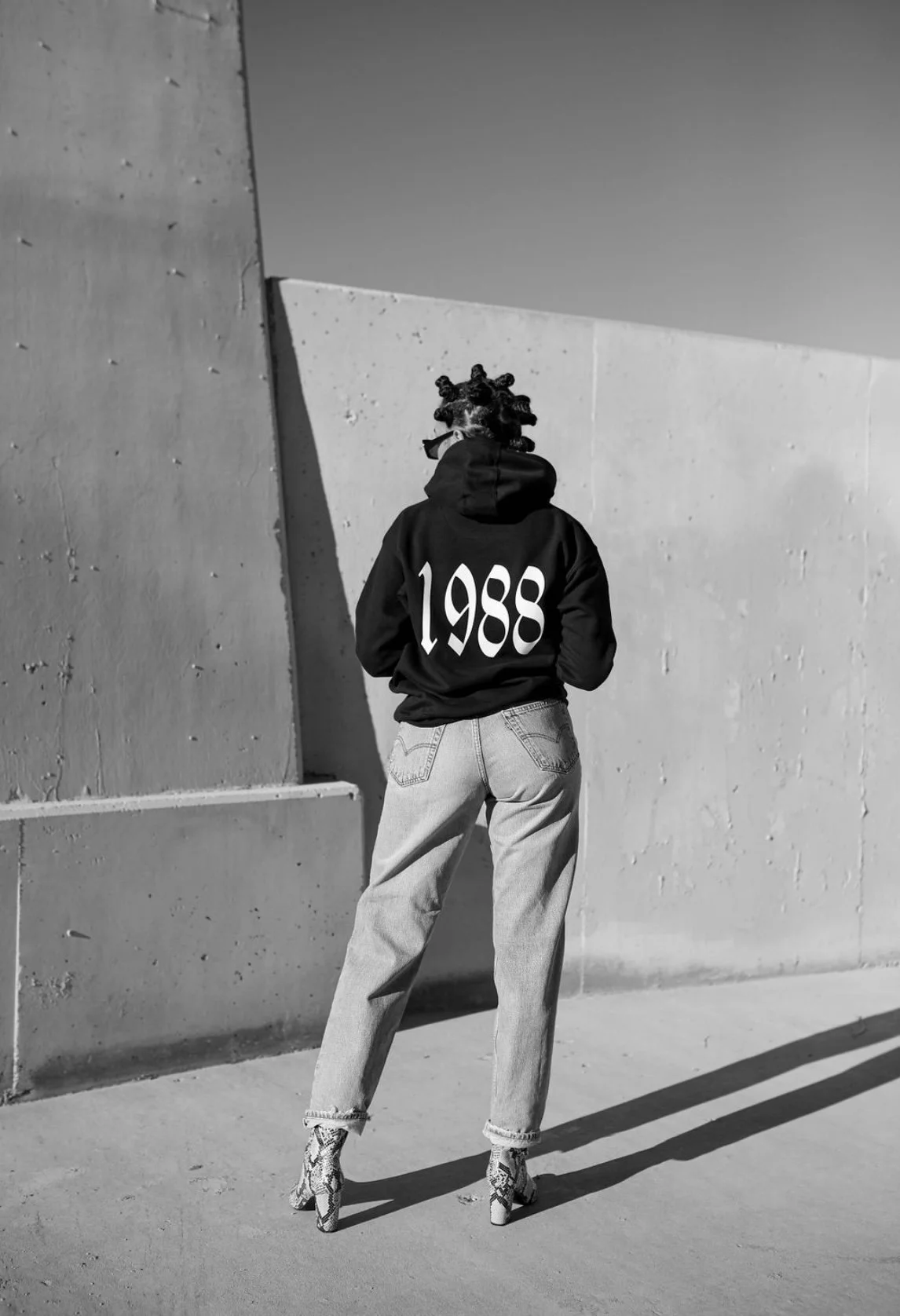 Black and white image of a person with short twisted hairstyle wearing a hoodie with '1988' on the back, light jeans, and high-heeled boots, standing against a concrete wall.