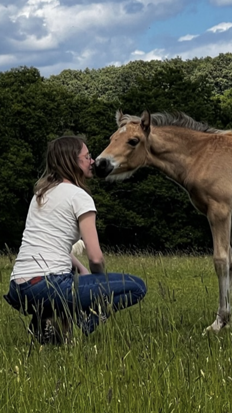 Rebecca, woman in her early forties crouches down in a green field wearing a white t-shirt and blue jeans. She is side-on to the viewer, eyes closed. A 6 week old bucksin foal strechs out to her and the foal and Rebecca touch noses. The sky is blue