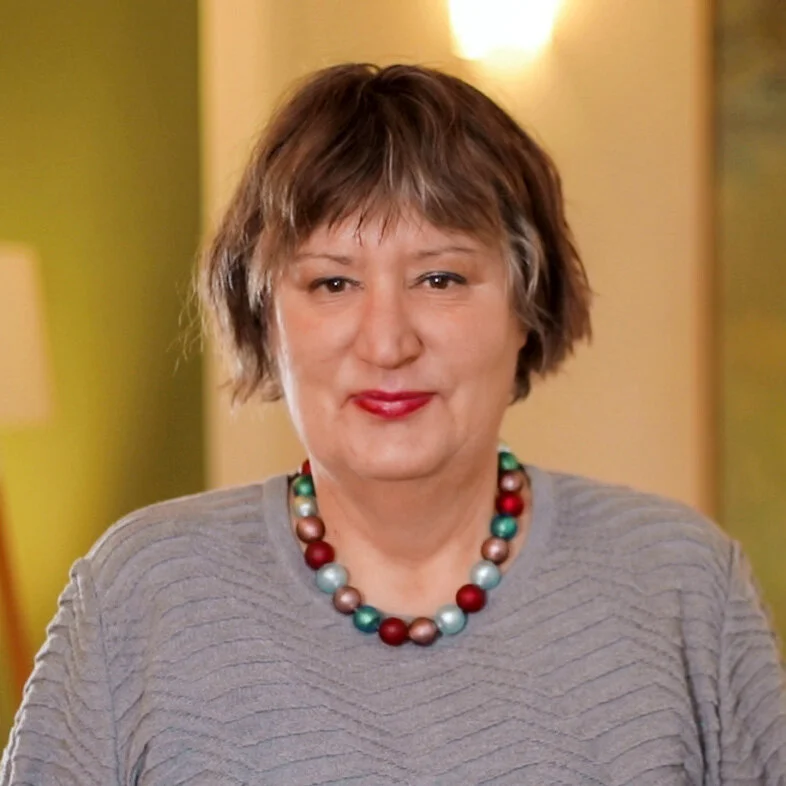 Nahaufnahme einer Frau mit kurzen braunen Haaren, rotem Lippenstift, grauem textured Oberteil und bunter Perlenkette, vor gelb-grünem Hintergrund.