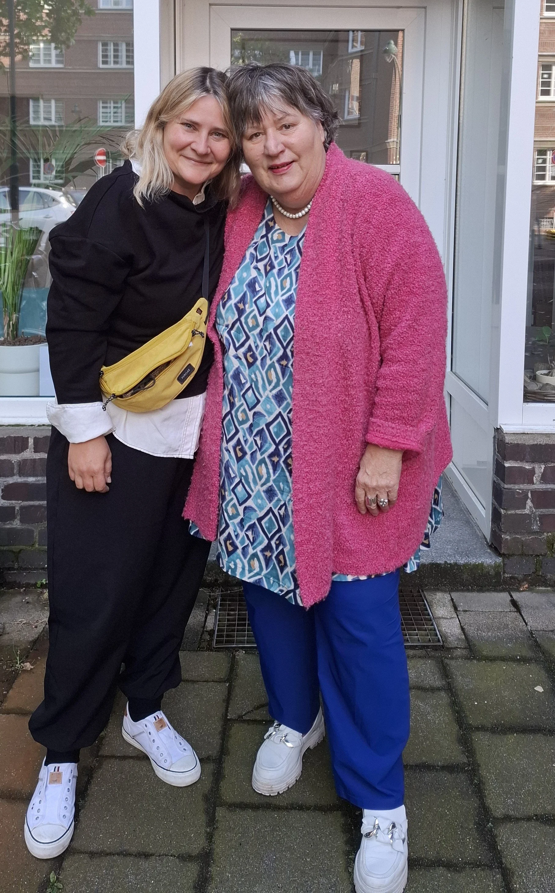 Ute Preiss und Christel van Dieken stehen zusammen vor einem Fenster, lächeln, eine trägt eine schwarze Jacke und eine gelbe Gürteltasche, die andere trägt eine pinke Jacke und eine blaue Kleid mit weißen Turnschuhen.