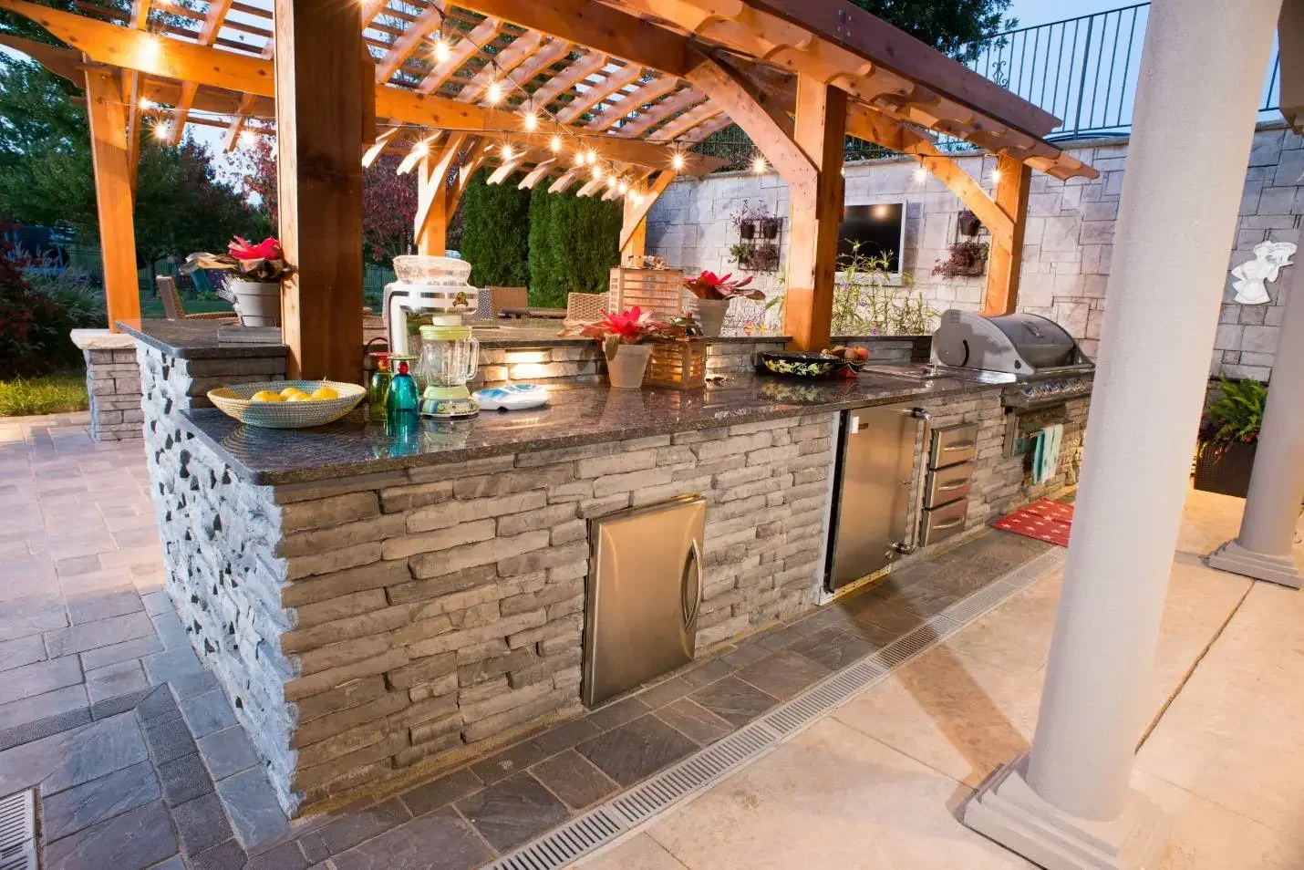 Outdoor kitchen with granite countertop, built-in stainless appliances, and string lights under arched cedar pergola at dusk at The Craftsman's Retreat by Green View in Illinois