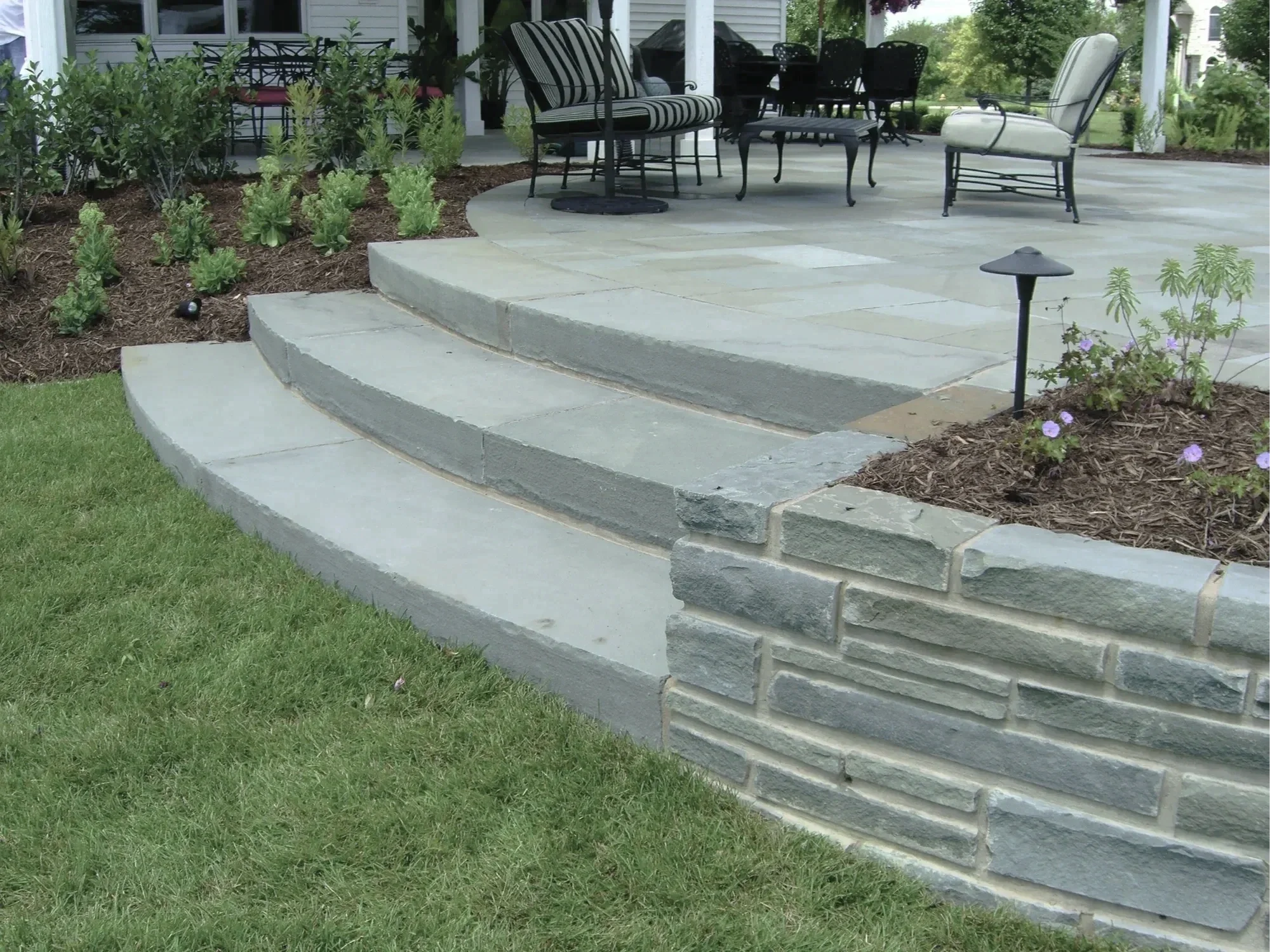 A backyard patio with stone steps leading up to a sitting area with outdoor furniture, surrounded by garden beds with plants and flowers, and a lamp post.