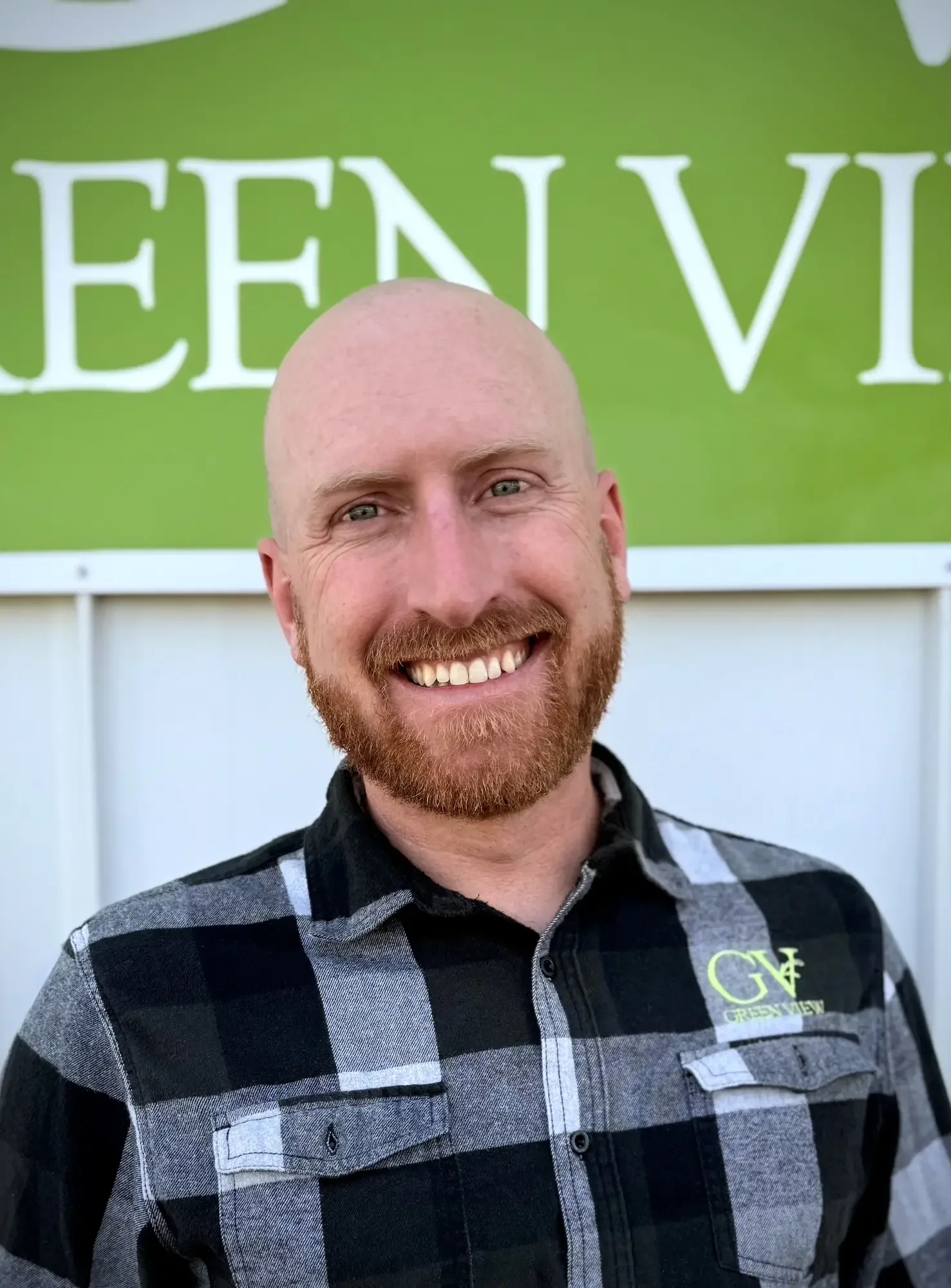 A smiling man with a red beard and shaved head, wearing a black and gray plaid shirt, standing in front of a green background with partial white text.