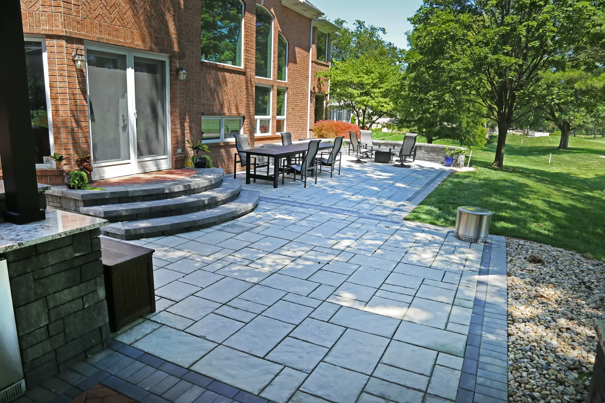 Wide large-format paver patio with outdoor dining, curved entry steps, and stone seating wall at The Wooded Pavilion by Green View in Illinois