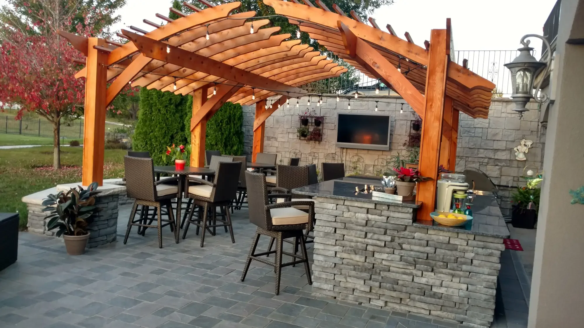 Arched cedar pergola with stone bar, wicker bar seating, outdoor dining, and stone TV wall with fireplace at dusk at The Craftsman's Retreat by Green View in Illinois