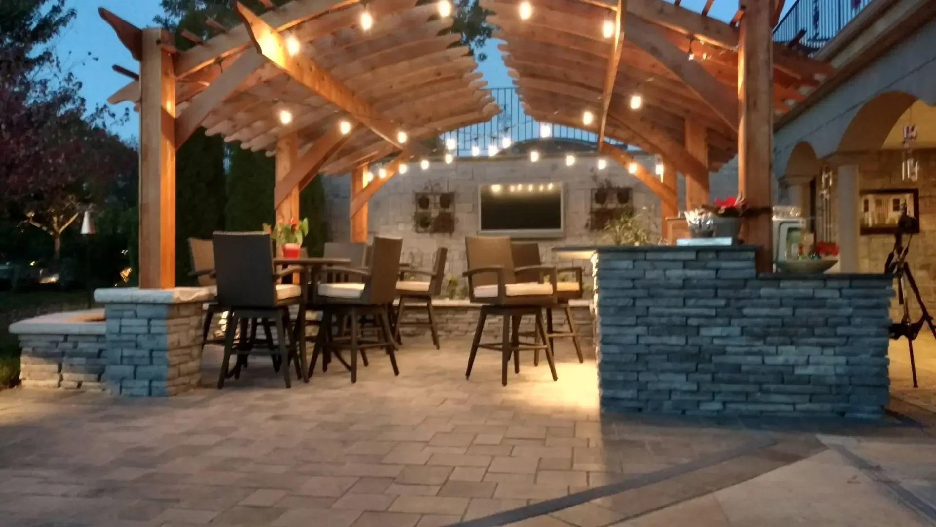Evening view of arched cedar pergola with string lights, stone bar seating area, and outdoor TV wall at The Craftsman's Retreat by Green View in Illinois