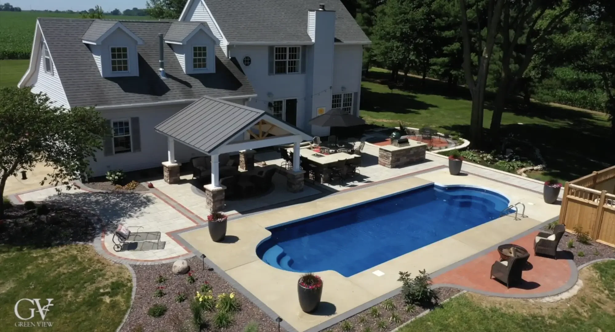 Aerial view of metal roof pavilion, pool surround, outdoor kitchen, and paver patio with red accent border at The Prairie Pavilion by Green View in Illinois