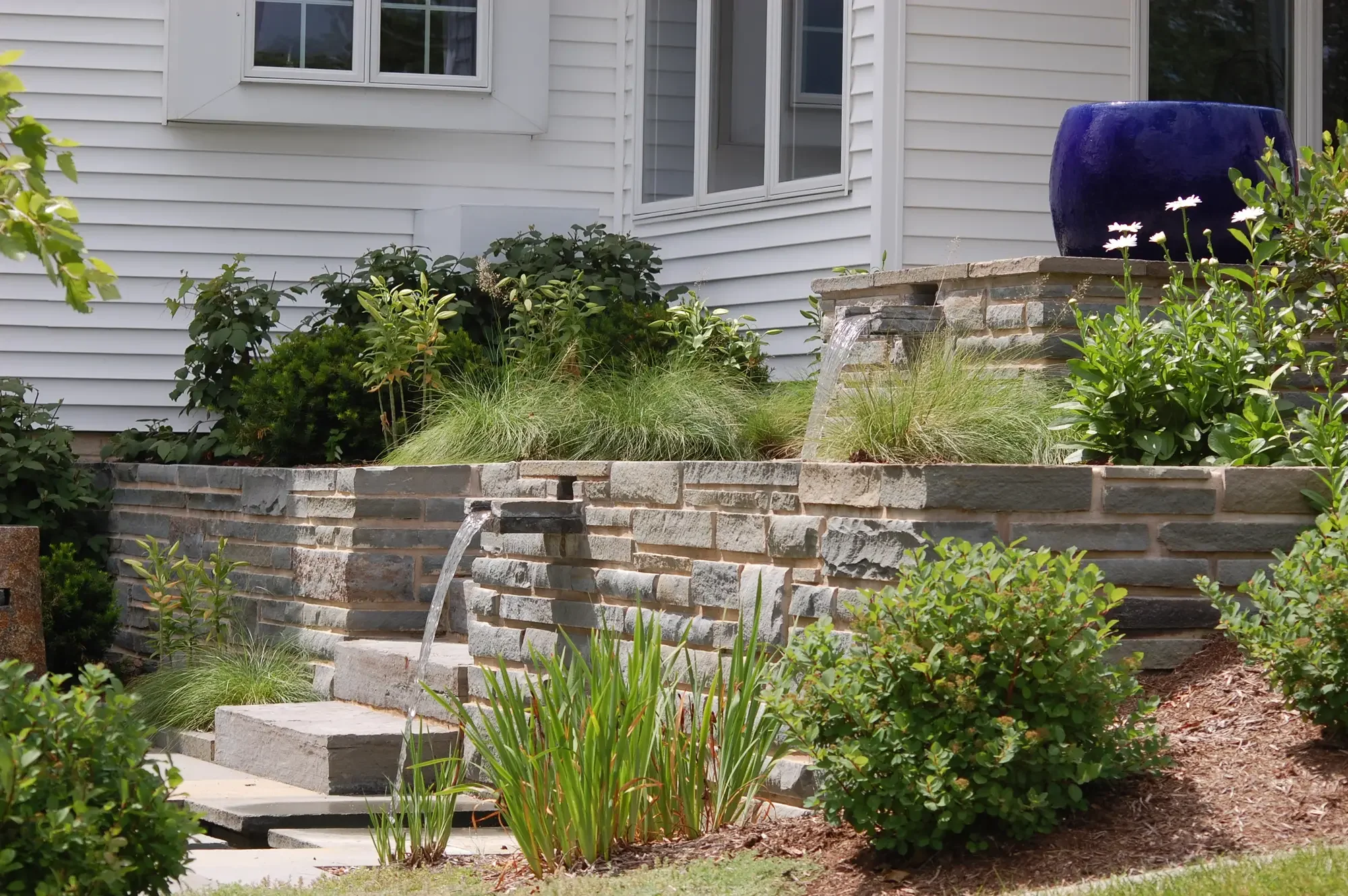 Stacked stone retaining wall with built-in water feature spouts, cobalt blue planter, and perennial planting beds at Harmony in Stone and Water by Green View in Illinois