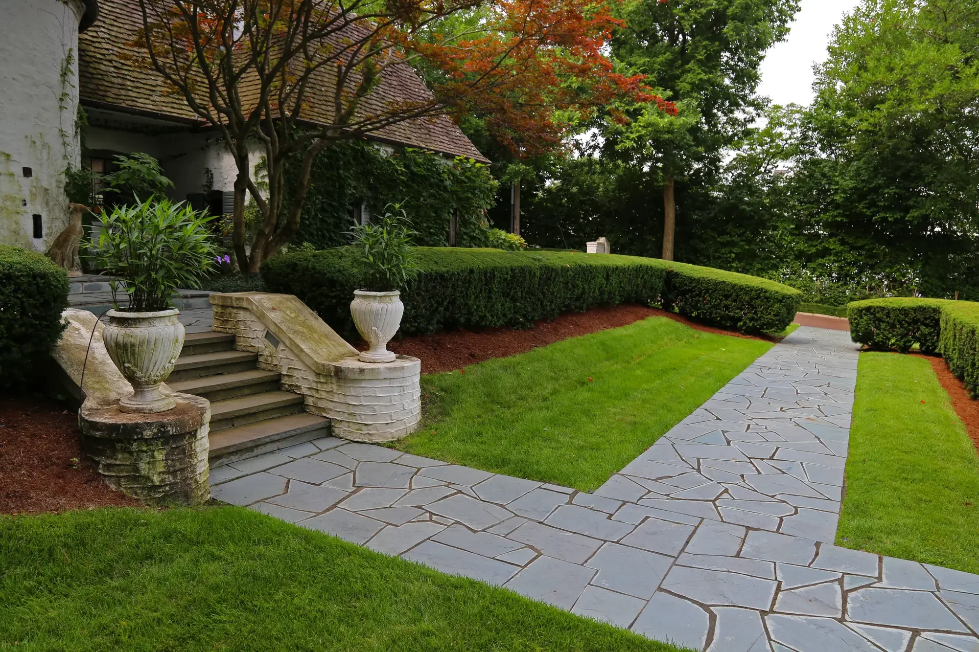 Flagstone walk with stone urn planters, curved clipped hedges, and garden gate entry at The Tudor Knot Garden by Green View in Illinois