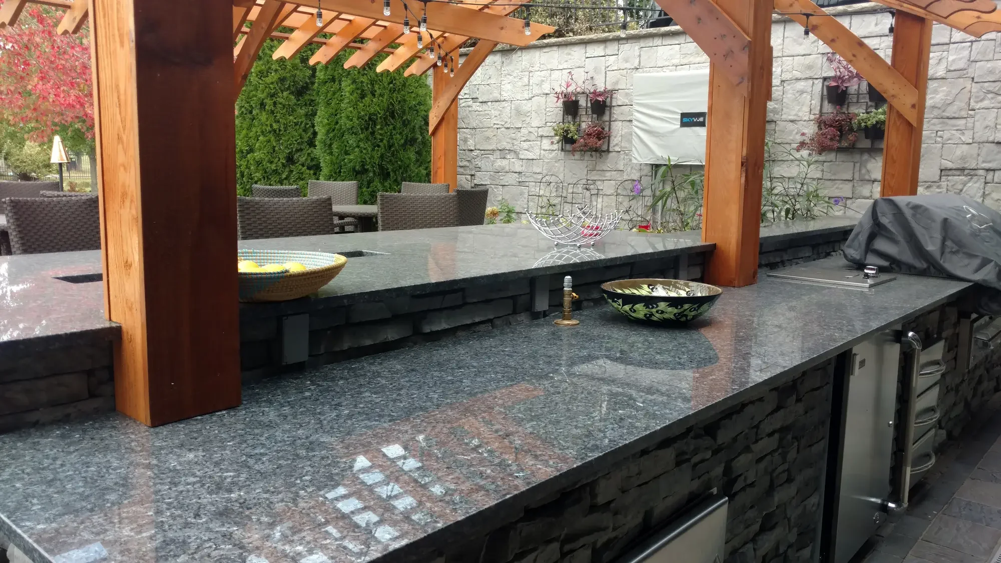 Outdoor kitchen with a granite countertop, wooden pergola, hanging lights, and garden seating area with trees and flowering plants in the background.